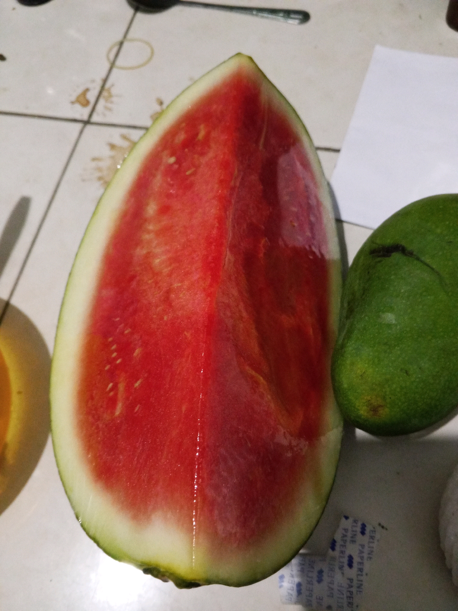 A slice of watermelon is placed on a tiled surface next to a green mango. The watermelon slice is a vibrant red with a white rind. The mango is oval-shaped and has a dark line across its surface. The tiled surface appears to be dirty with some smudges and a rubber band. A piece of white paper is visible in the background. The scene appears to be indoors, possibly in a kitchen or dining area. The lighting suggests it is daytime, with artificial light also contributing. The overall impression is one of a casual setting where fruit has been prepared or is about to be consumed. The specific location in Karawang, Indonesia, cannot be determined from the visual information provided in the image.