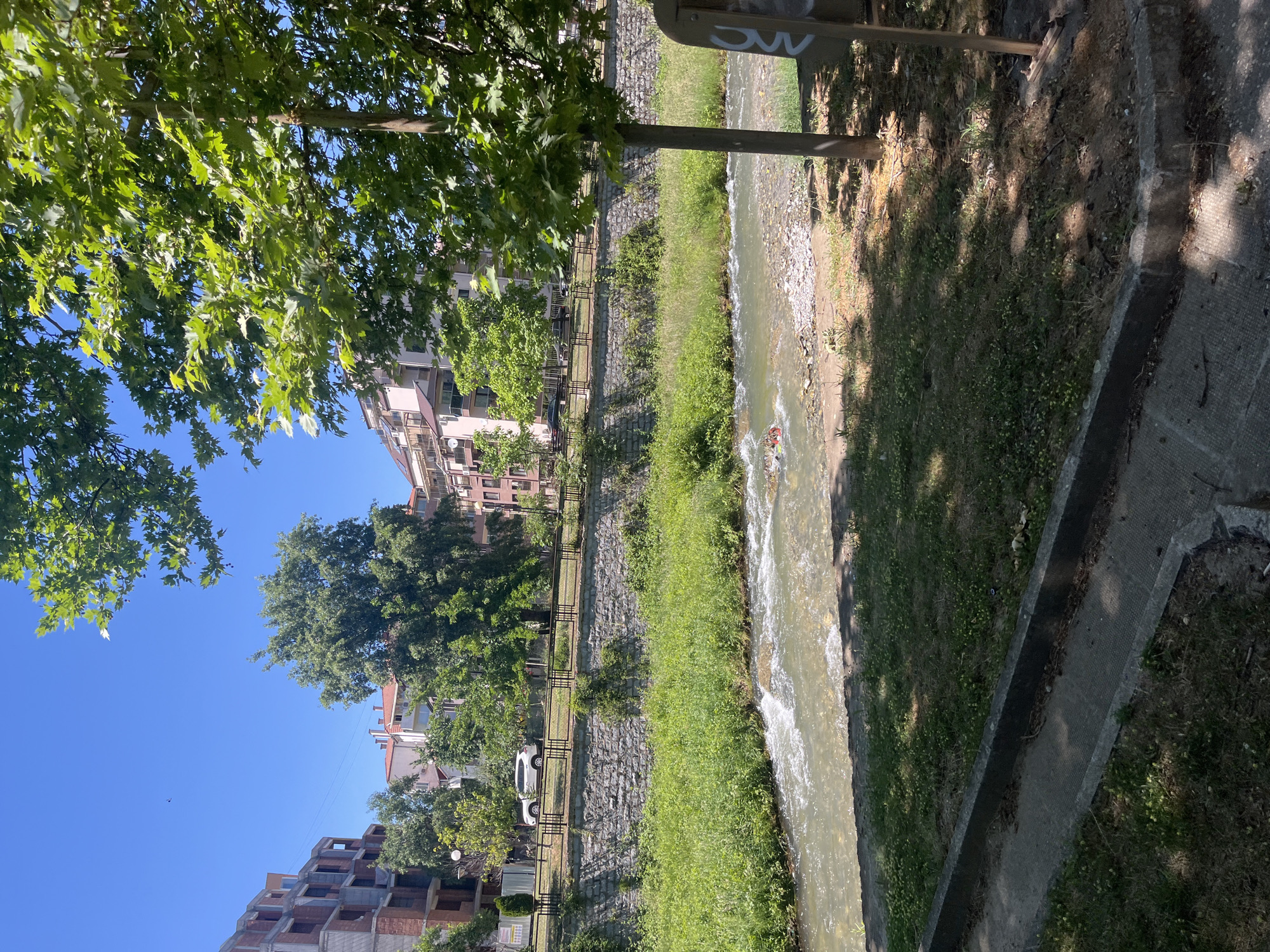 The image shows a waterway running through an urban area. The buildings located in the background suggest this might be in the city of Blagoevgrad, Bulgaria. A narrow stream of water flows from the top to the bottom of the frame, flanked on either side by grassy banks. The grassy banks are separated from the walkway with low stone walls and dark metal railings. On the walkway side is a single white car. Overhanging foliage from trees casts shadows on the concrete pathway and the grassy areas. A metal pole with graffiti writing on it is visible in the top right corner. The scene conveys a sense of urban nature integration.