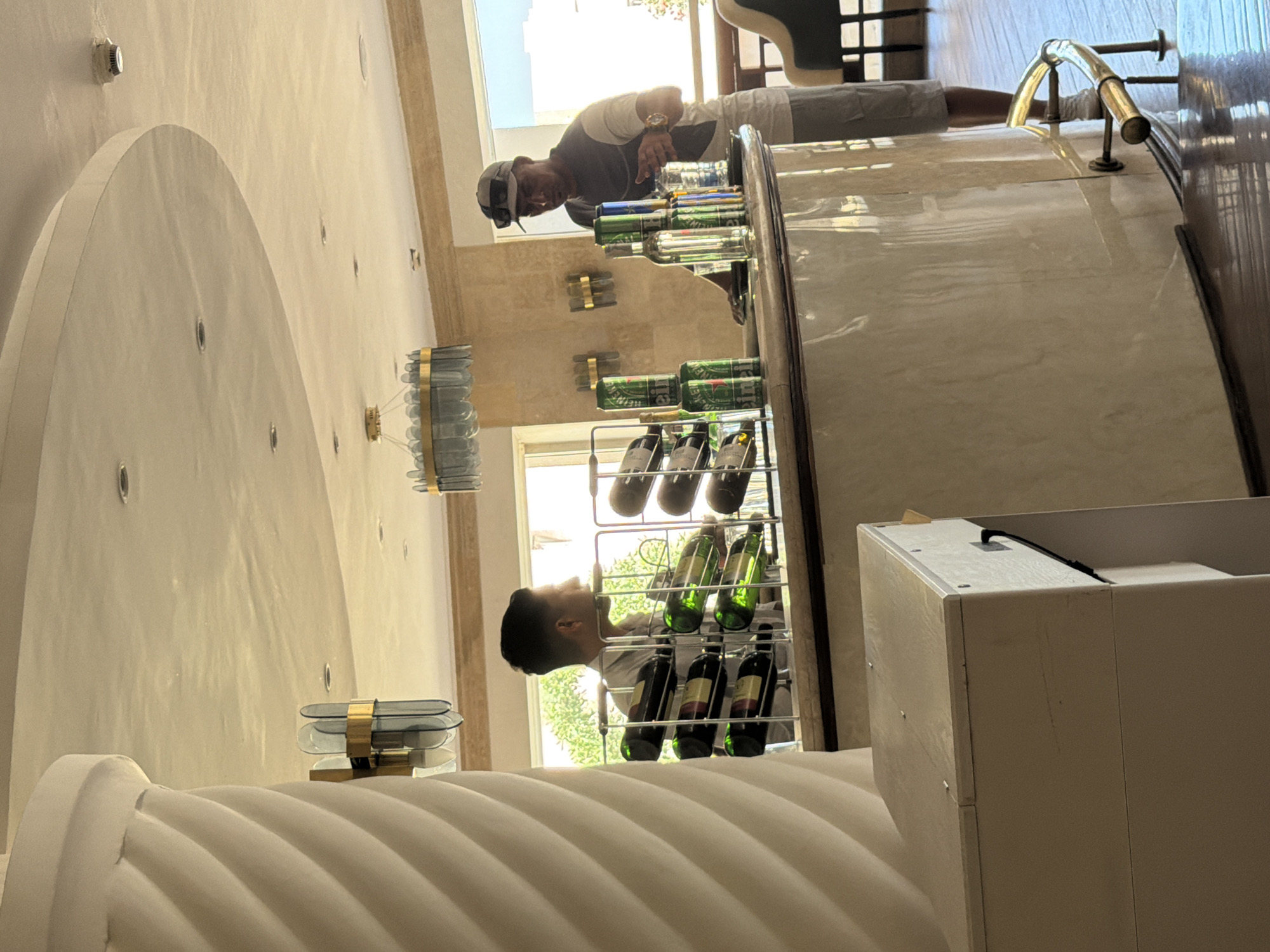 The image captures an indoor bar scene, likely within a hotel in Sharm El Sheikh, Egypt, given the architectural style and lighting. On the left, a man in a light-colored shirt is looking towards the right, positioned behind a rack of wine bottles. In the center and right, a man wearing a baseball cap, sunglasses, and a two-toned t-shirt stands behind the bar. He is in the process of serving or preparing drinks, with several bottles of alcohol and cans of Heineken visible on the bar top. The bar itself is made of a light-colored, reflective material. The background reveals large windows with natural light streaming in, showing greenery outside. The ceiling has recessed lighting and a decorative chandelier. A decorative column is visible on the far left.