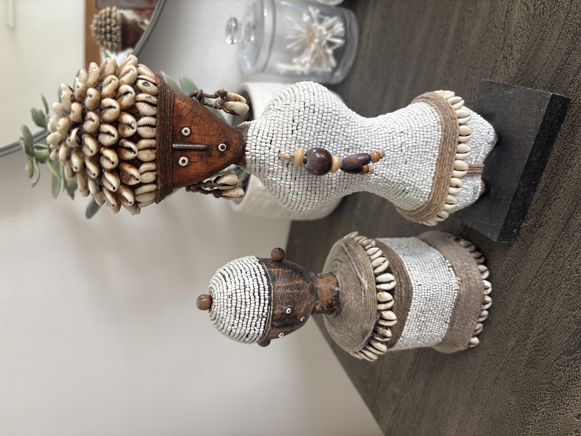 Two handcrafted figures adorn a dark wooden surface. The figure on the left is shorter and wears a white beaded cap topped with a wooden bead. Its body is covered in white beads, with a band of twine and shells around its middle and base. The figure on the right is taller, with a headdress of numerous small shells. Its face is carved from wood and adorned with white bead eyes and mouth, and two wooden beads at the sides of its head. Dangles of shells hang from its ears. Its body is covered in white beads, with a band of twine and shells around its waist, and a beaded pendant hanging from its chest. The background features a potted plant and a glass jar filled with cotton swabs. The overall scene is indoors, suggesting a decorative display. There is no indication that this scene is located in Dubai, United Arab Emirates.