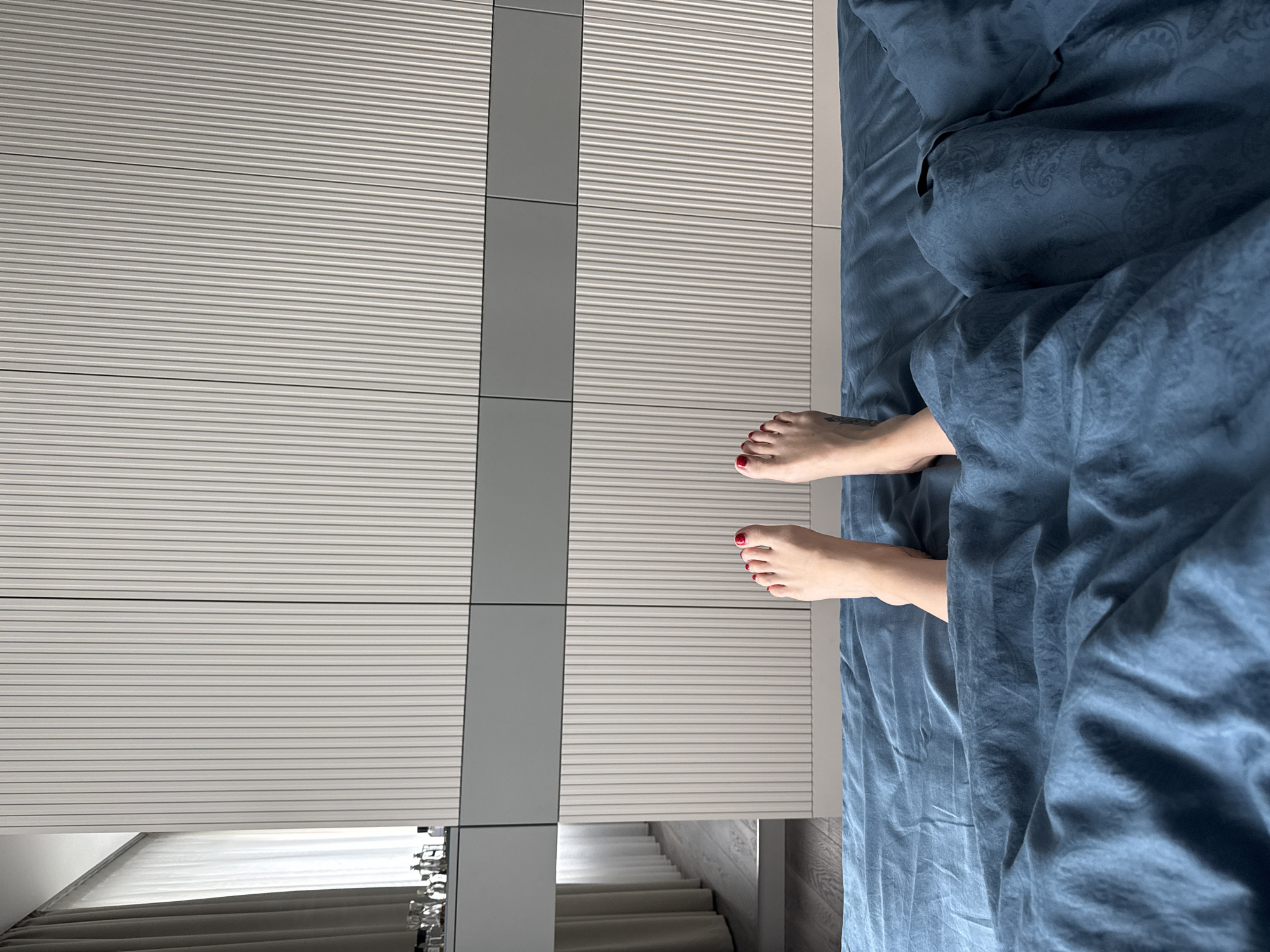 A top-down shot shows the feet of a person with red nail polish sticking out from under a blue patterned duvet. The person is presumably in bed. Beside the bed is a wall or wardrobe with a horizontally striped texture. The location is inferred to be Khimki, Russia, based on prior information, although there is no visual evidence in the image to confirm this. There is a reflection of a staircase in the wall.