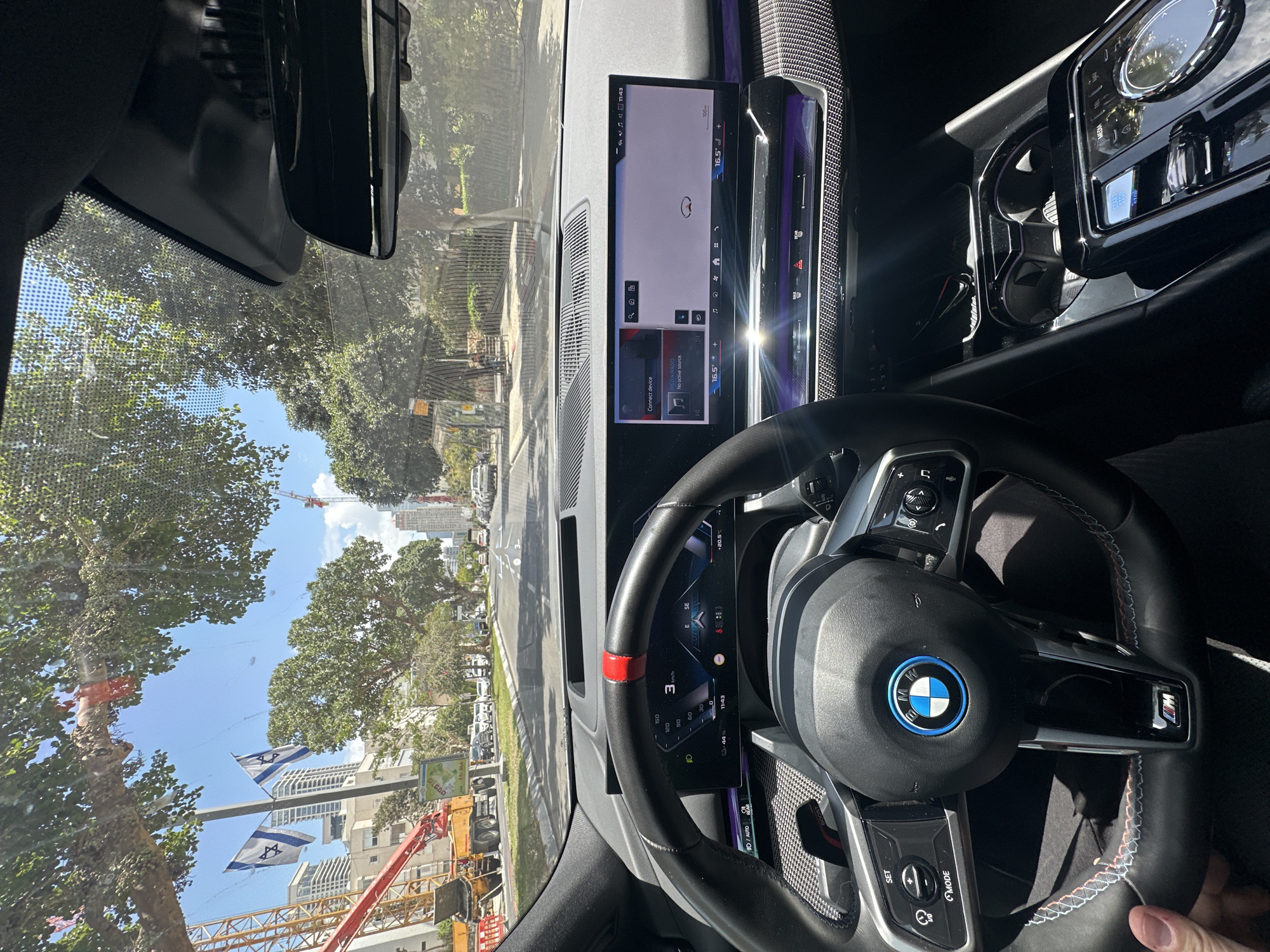 The image is taken from inside a BMW vehicle, showcasing the dashboard, steering wheel, and infotainment screen. The navigation system on the infotainment screen is active. Outside the car's windshield, the view shows a street scene in Tel Aviv, Israel, identifiable by the presence of Israeli flags. The street has buildings, trees, and construction cranes in the background. The sky is clear and blue. The steering wheel is black leather with a red marking at the top. Various buttons and the BMW logo are visible on the steering wheel.
