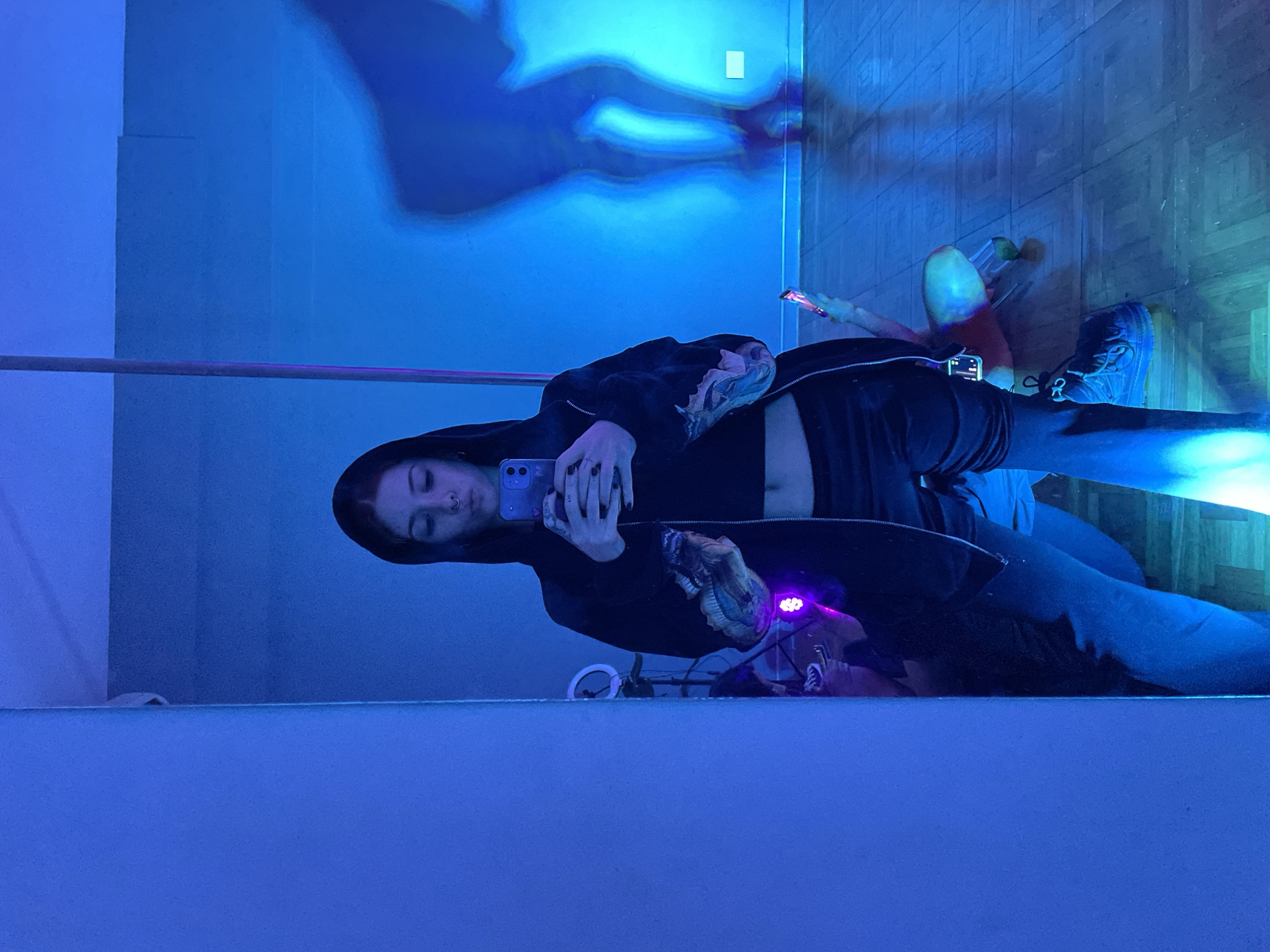 A young woman is taking a selfie in a mirror. She is wearing a black hooded jacket over a black crop top, blue jeans, and blue sneakers. She is holding a blue smartphone. The room is lit with blue lighting. There is a ring light in the mirror. The floor appears to be hardwood. Based on visual elements, the location is not determinable to be Buenos Aires, Argentina.