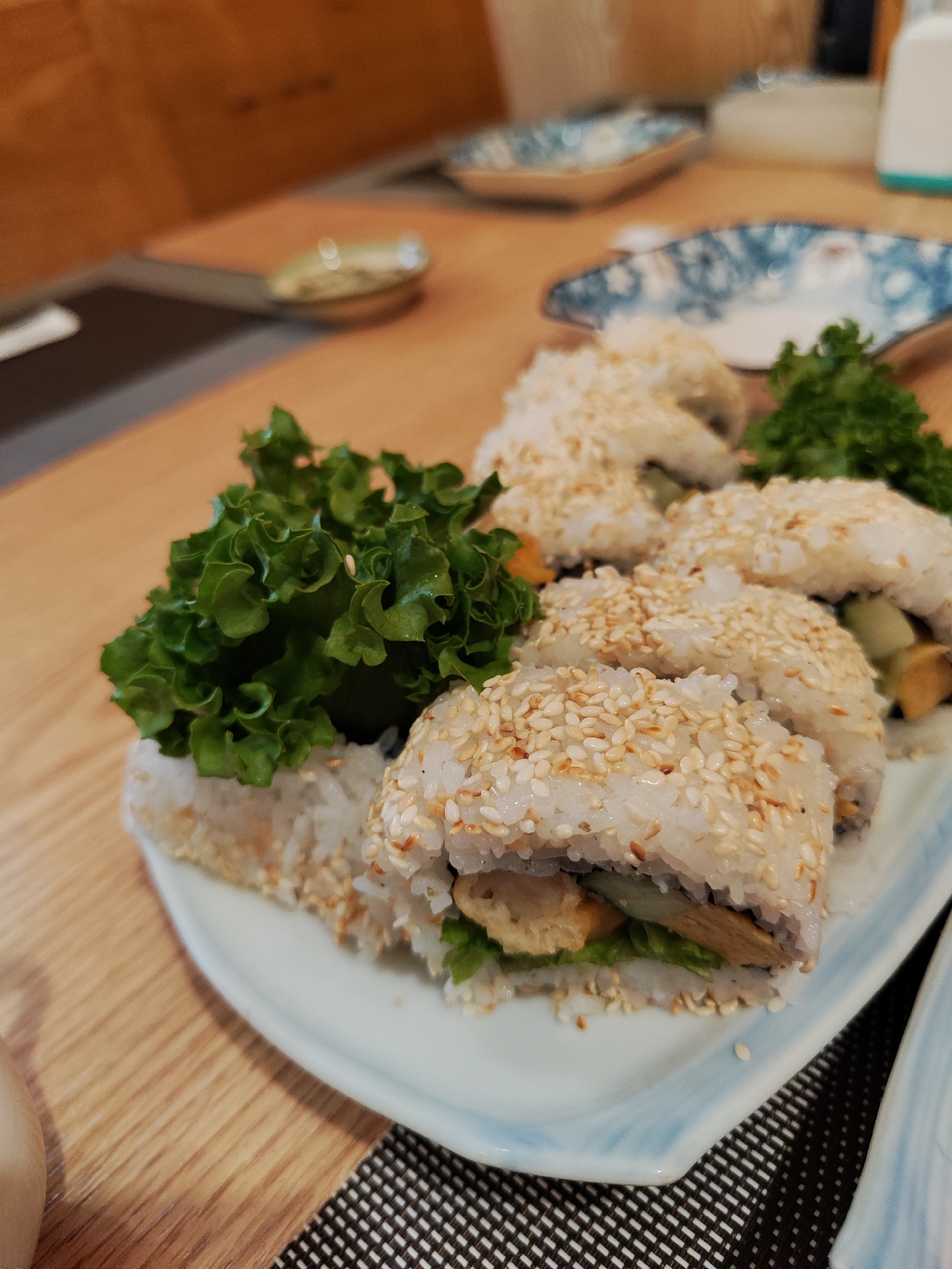 The image shows a plate of sushi rolls sprinkled with sesame seeds, garnished with lettuce, on a table covered with a black and white checkered placemat. The table has a light wooden surface. In the background, there are various small plates and a wooden wall. The image was taken in Da Lat, Vietnam.