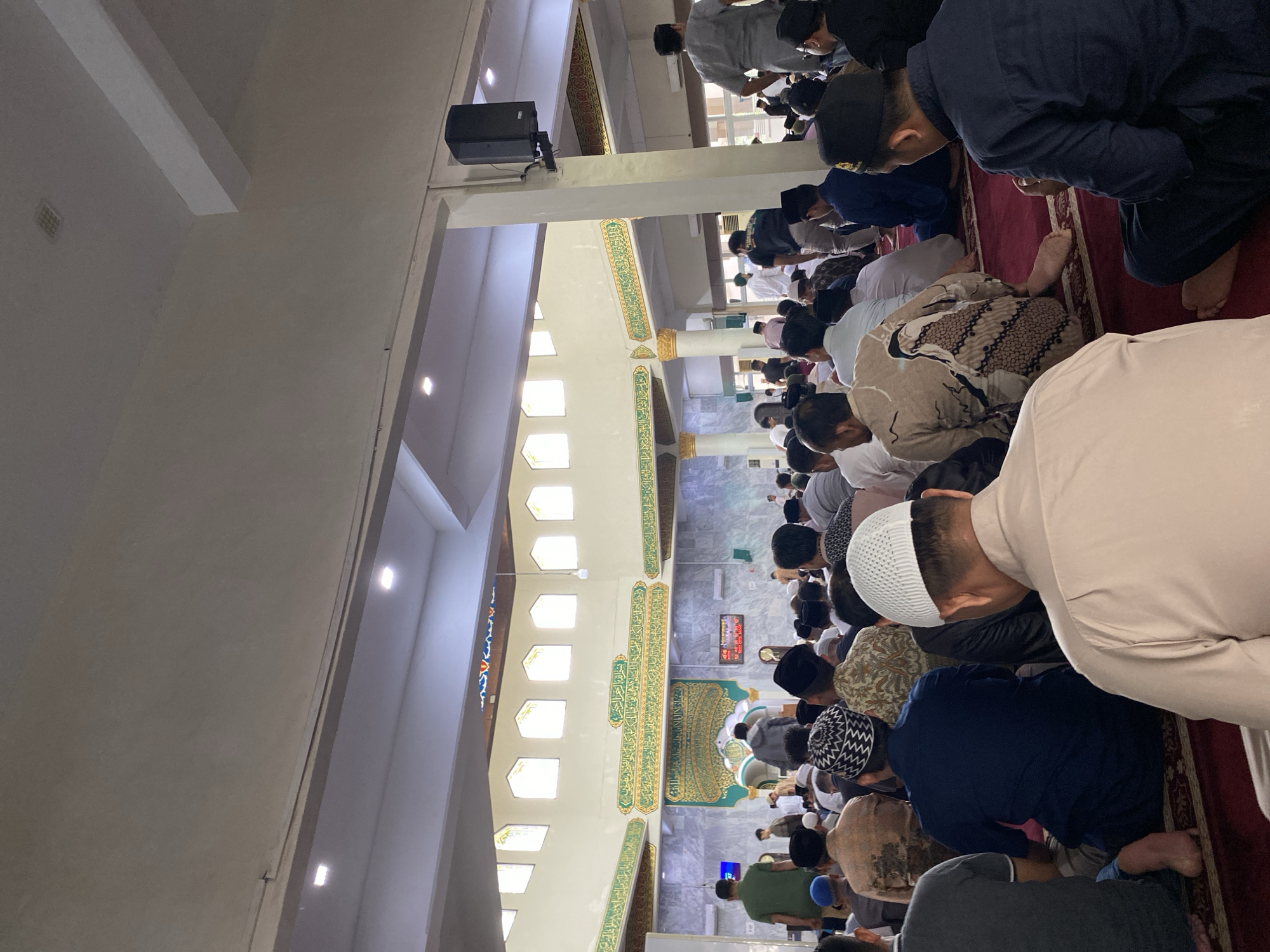 The image depicts a group of men engaged in prayer, likely within a mosque. The scene is oriented vertically. The men are arranged in rows, facing the same direction, and are in the process of performing the Ruku' (bowing) posture of Islamic prayer. They are dressed in various traditional Muslim attire, including prayer caps (peci) and long shirts or robes. The floor is covered with a red carpet, typical of prayer spaces.
In the background, architectural elements of the building are visible, including white walls, columns, and decorative friezes with Arabic calligraphy in green and gold. Several rectangular windows are present, letting in natural light. A black speaker is mounted on a pillar in the upper left portion of the image, and another digital display is visible in the distance. The overall atmosphere appears solemn and devout. Given the context of Lhokseumawe, Indonesia, this is likely a congregational prayer, possibly Zuhur or Asr prayer as it is daytime. No specific weather conditions are discernible. The lighting suggests it is daytime.