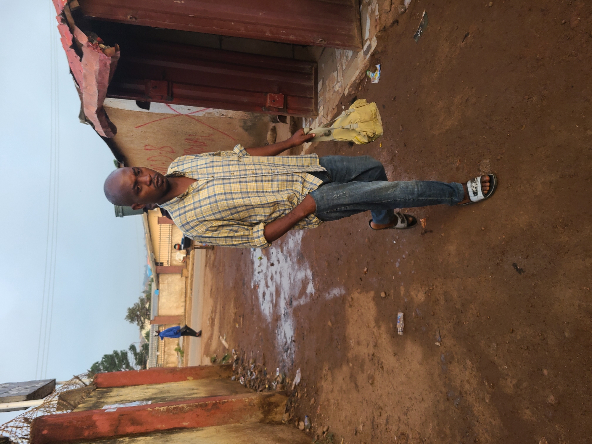A man stands on a dirt path in Jos, Nigeria. He has a shaved head, wears a yellow plaid shirt, jeans, and sandals. He is holding a yellow bag or bundle. The path is wet in places, with trash and debris along the edges. Buildings border the path, one with a red metal door. Another person walks in the background.