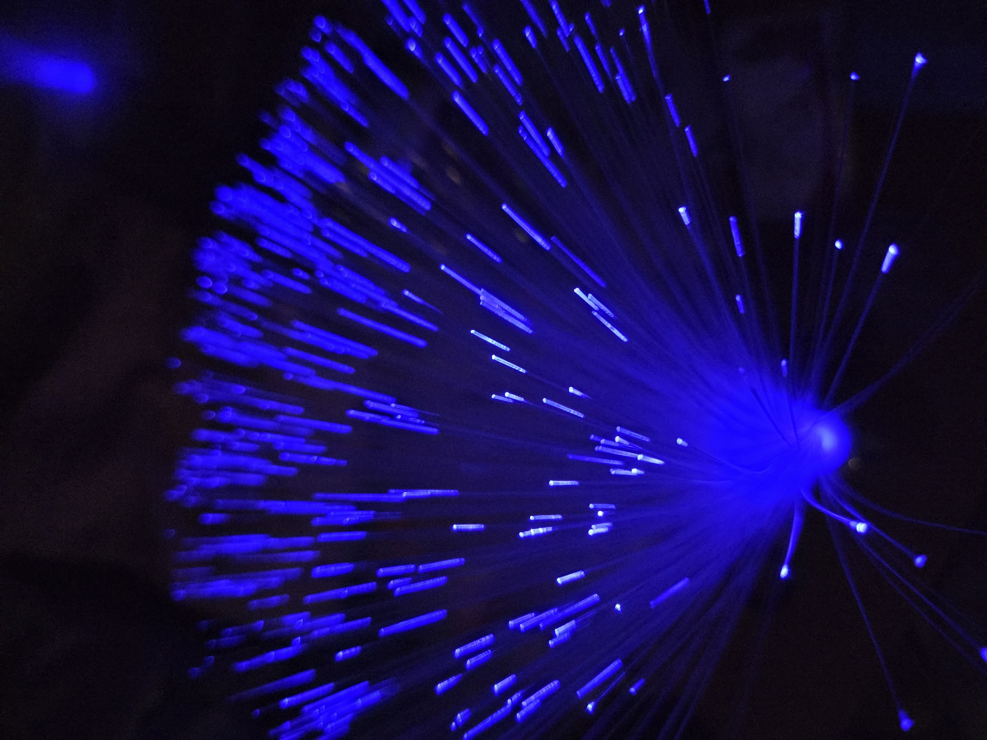A close-up shot shows a fiber optic lamp emanating blue light. The light sources are arranged in a radial pattern from the base and are set against a dark background. No people are visible in the image. The location is not discernible from the visual information.