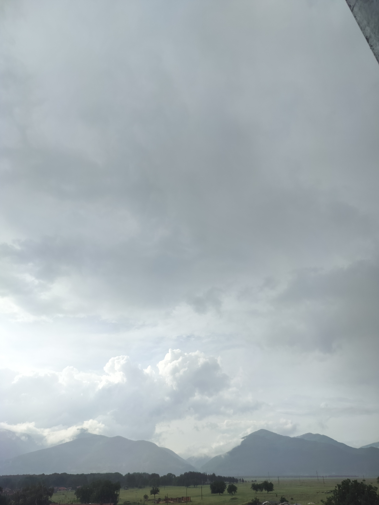 The image captures a wide, scenic landscape under a heavily overcast sky. The upper two-thirds of the frame are dominated by dense, gray and white clouds, with brighter, fluffier formations visible in the lower left, suggesting the sun attempting to break through.
In the middle ground, several layers of mountains extend across the horizon, appearing hazy and muted in shades of blue-gray due to atmospheric conditions and cloud cover. Below the mountains, a dark, horizontal strip of dense trees, likely a forest, stretches across the landscape. The foreground features a vast, open green field or pasture. Scattered darker trees are also visible within this field, and faint hints of utility poles or lines can be discerned further back in the field area. On the very bottom left, small structures, possibly houses or farm buildings, are partially visible.
The scene depicts a calm, natural setting, with no human or animal activity overtly visible. The weather is distinctly cloudy and overcast, creating a diffused light that suggests either an early morning or late afternoon with heavy cloud cover, or a generally dull day. The overall impression is one of serene, rural natural beauty in a mountainous region, consistent with a location like Gorno Sahrane, Bulgaria. No text is visible in the image.