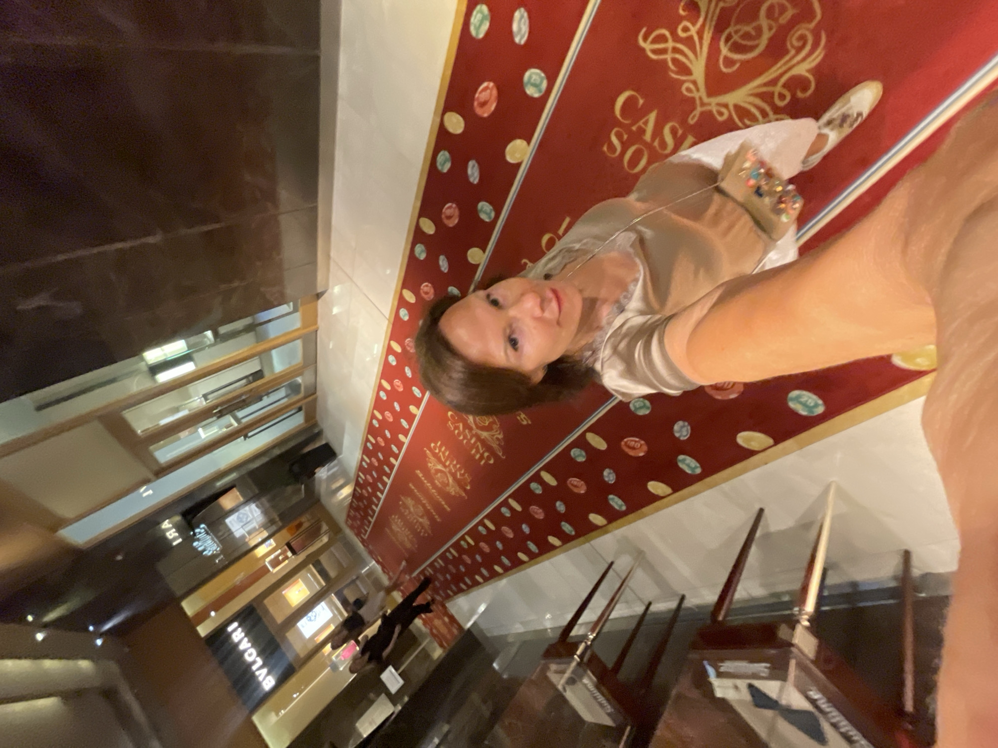 In the city of Estosadok, Russia, a woman takes a selfie. She is standing on a red carpet in what appears to be a casino or a high-end establishment. The carpet is decorated with colorful circles resembling casino chips and a golden crest. To her left, a store with the "BVLGARI" logo is visible. The woman is wearing a shimmering beige top, white pants, and sneakers. She is holding a small, beaded clutch purse. In the background, two other individuals are walking away from the camera. The environment is indoors, with polished floors and reflective surfaces.