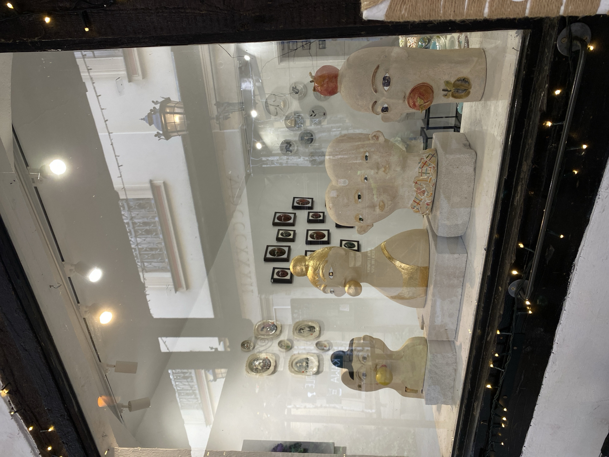 This image captures a street-level view through the display window of an art gallery or shop in Wrocław, Poland. Four distinct, anthropomorphic sculptures, likely ceramic or clay, are prominently displayed on individual light grey rectangular plinths within the window.
From left to right, the sculptures are:
1. A light-colored bust with an exaggerated face, a blue cap-like element on its head, and a yellow spherical object protruding from its mouth.
2. A light-colored bust featuring a golden topknot and a golden band across its chest, with a golden spherical object in its mouth. Its eyes appear closed or stylized.
3. A wider bust depicting two conjoined heads or faces, with multiple eyes and a distinct, multi-colored patterned collar composed of small tesserae.
4. An elongated, light-colored head-like bust with an open mouth from which a red fruit, possibly an apple, protrudes. A smaller, darker fruit motif is visible on its lower cheek, and a red apple balances on top of its head.
Behind the sculptures, various artworks are visible on the interior walls. To the left, a collection of oval and circular framed pieces hangs, while to the right, a grid of dark-framed rectangular artworks, each containing a dark, oval object, is displayed. Further to the right, a series of circular framed pieces with distinct designs is seen. The interior is illuminated by track lighting with several spotlights.
The glass of the display window exhibits significant reflections, revealing elements of the urban environment across the street. A light-colored building with dark, ornate balconies and windows is clearly reflected, along with a prominent, dark, decorative street lantern featuring illuminated glass. A string of small, warm-toned LED lights is strung along the dark, possibly wooden, lower frame of the display window, adding a decorative element to the foreground. The window itself shows minor smudges and reflections consistent with a public street-facing frontage.