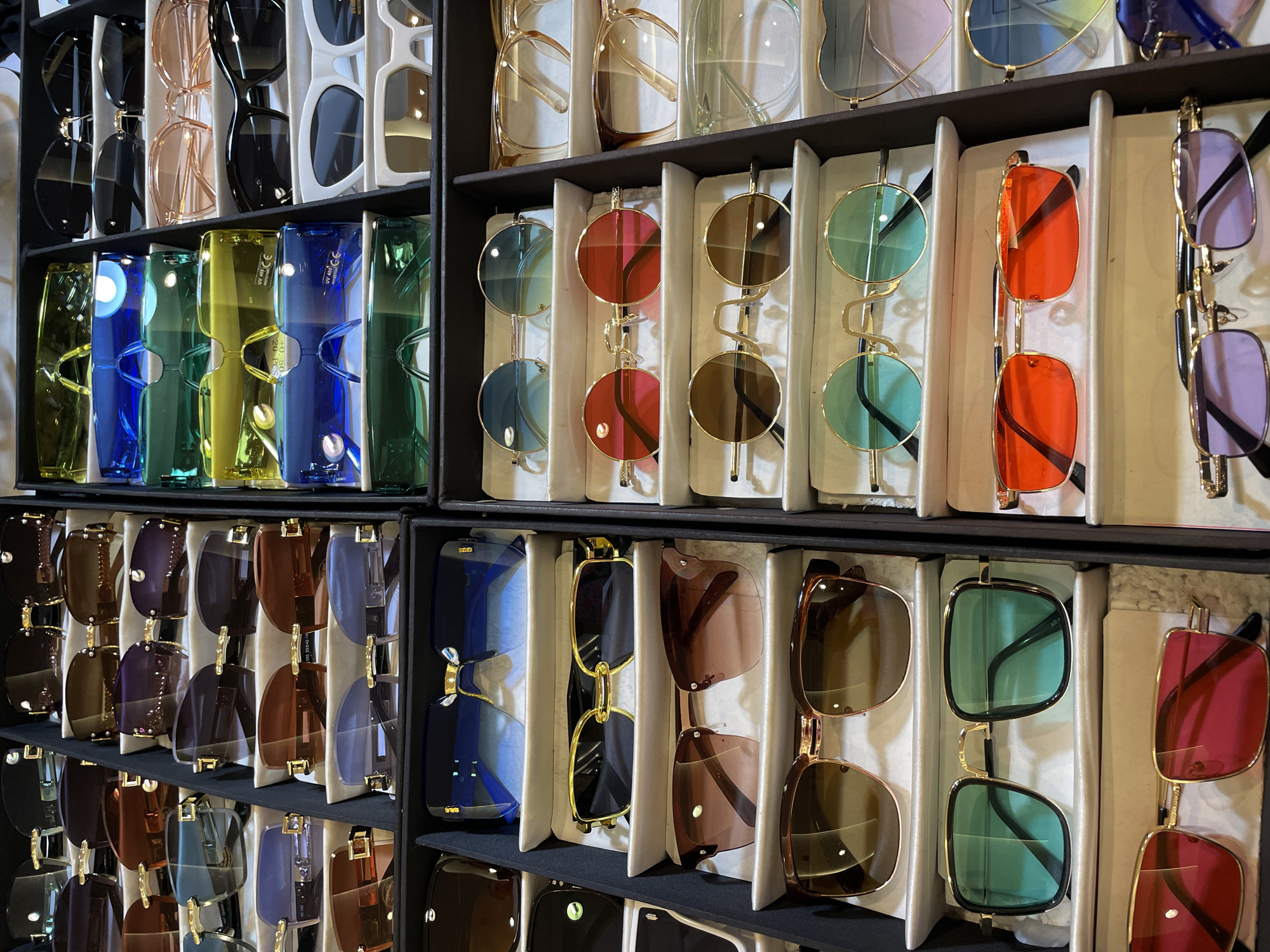 The image shows a display of various sunglasses in a store. The sunglasses are arranged in rows and compartments. There are different styles, colors, and lens tints visible, including aviator, round, and square frames, with lenses in blue, green, red, pink, yellow, gradient, and mirrored finishes. The store appears to be a retail environment, possibly a market or a specialized sunglasses shop. The scene is indoors, with artificial lighting. No people are visible in the image. The specific location within Pattaya City, Thailand cannot be determined from the image alone.
