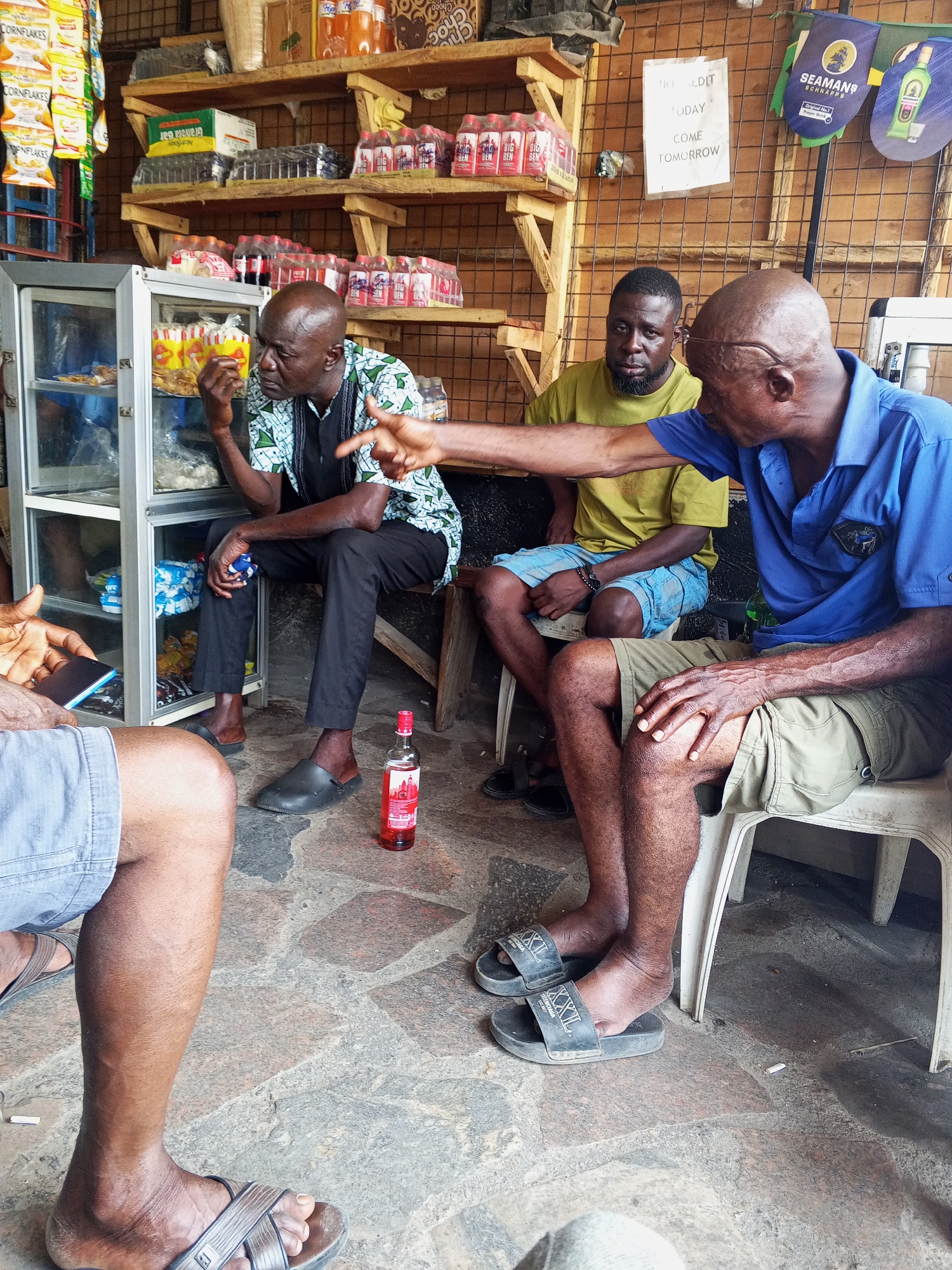 In a likely informal setting in Zango-ndaji, Nigeria, four African men are gathered. One man, in a blue shirt and khaki shorts, sits on a white plastic chair, gesturing with his right arm extended. To his left, another man in a yellow t-shirt and blue shorts sits on a stool. A third man, wearing a patterned shirt and black pants, sits on a wooden stool beside a glass-door display case stocked with food items; he holds something in his hand near his mouth. Only the legs of the fourth man are visible, clad in blue shorts and sandals. A bottle of liquor rests on the floor. In the background are shelves laden with various products, including drinks and boxed goods. A sign reads, "No Credit Today, Come Tomorrow." Advertising for "SEAMANS Schnapps" is also visible. The floor is composed of tiled stone.