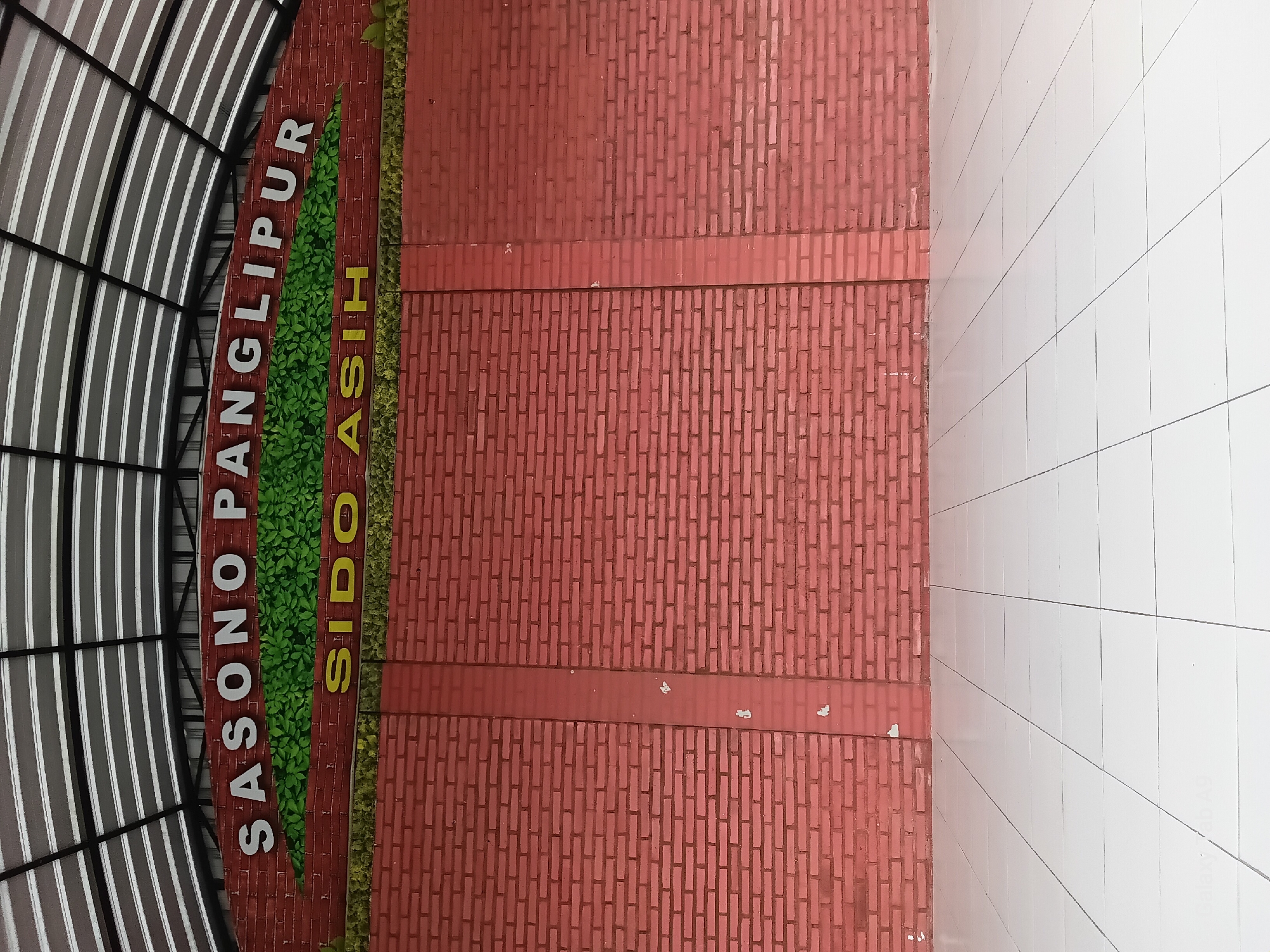 The image depicts the facade of a building with a red brick wall under a white, corrugated roof supported by metal beams. The building's name is displayed in large, white, raised letters on the upper portion of the brick wall, reading "SASONO PANGLIPUR," with a decorative band of green foliage underneath. Below this, a smaller sign features yellow raised letters that spell out "SIDO ASIH." The brick wall is segmented by a vertical strip of slightly darker red brick. The foreground shows a tiled floor, composed of large, white square tiles with thin black grout lines. The overall scene is outdoors, with no people or immediate activity visible. The architecture suggests a public space or pavilion, possibly in a park or community area.