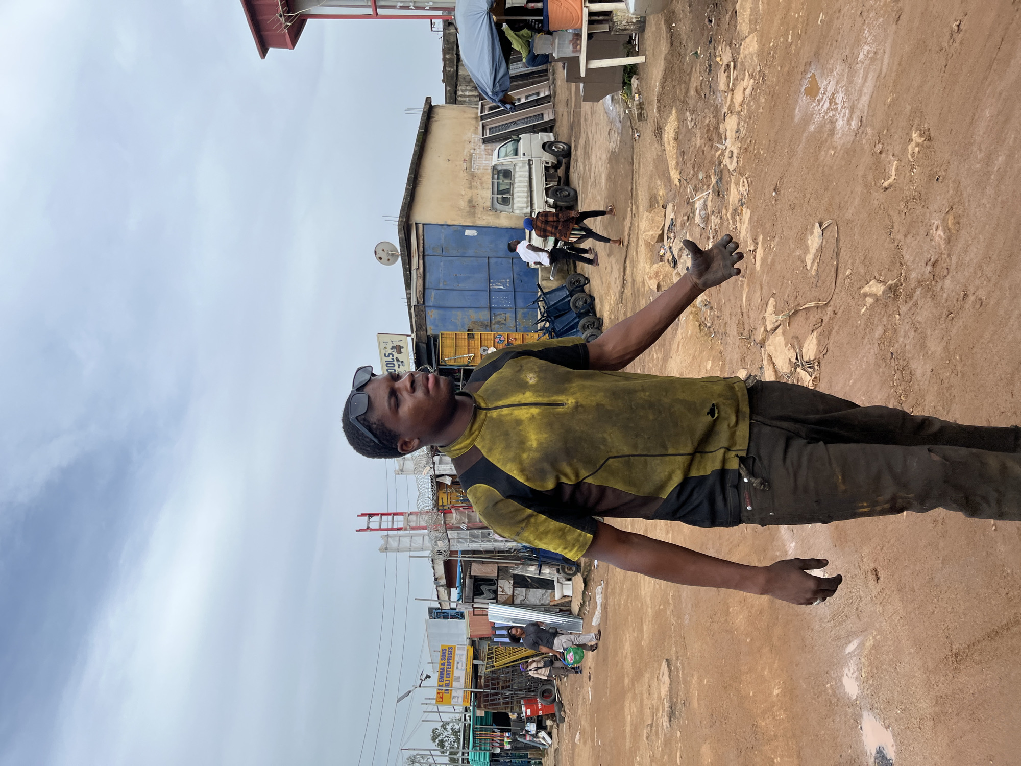 In Gura Top, Nigeria, a Black male, likely a construction worker, stands outdoors on a dirt road. He wears a dirt-stained yellow and black short-sleeved shirt with a zippered collar and dark pants that are also soiled. Sunglasses are perched on his head. His right arm is extended, with his hand open and covered in dirt. The background reveals a busy marketplace with various stalls and structures. A white truck is parked nearby, and several people are visible in the distance, engaged in their activities. The ground is uneven and dusty, with some puddles of water. The sky is overcast.