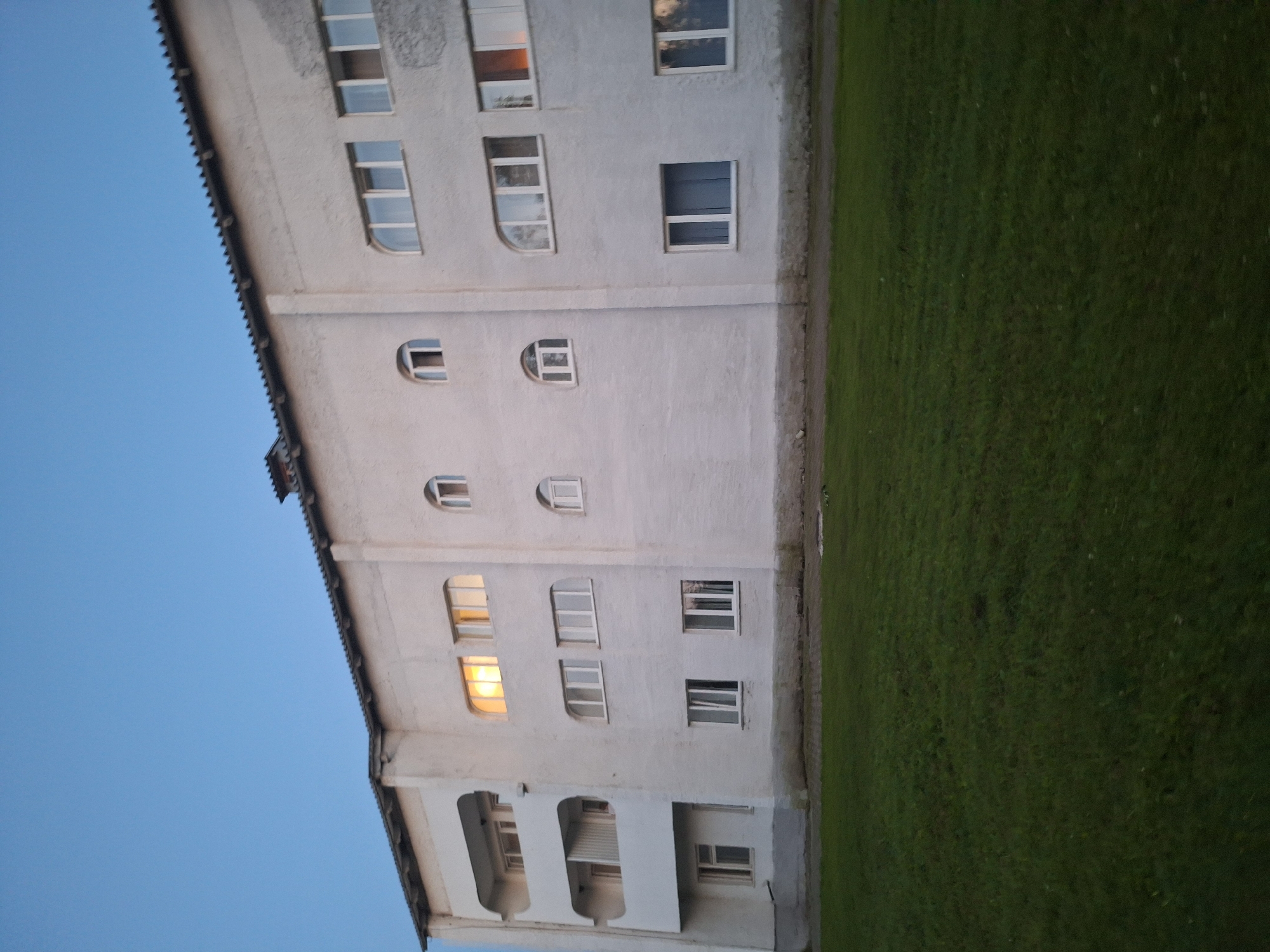 A low-angle outdoor shot captures the exterior of a multi-story apartment building in Atamanovka, Russia, under a clear, pale blue twilight sky. The building, painted white and showing signs of weathering, features several rows of windows and balconies. On the left side, there are three levels of recessed balconies. Various window shapes are visible, including rectangular, arched, and semi-circular designs. One set of upper-floor windows on the left side is illuminated, indicating interior lights are on. The building stands adjacent to a well-maintained grassy area. The overall scene depicts a quiet residential setting.