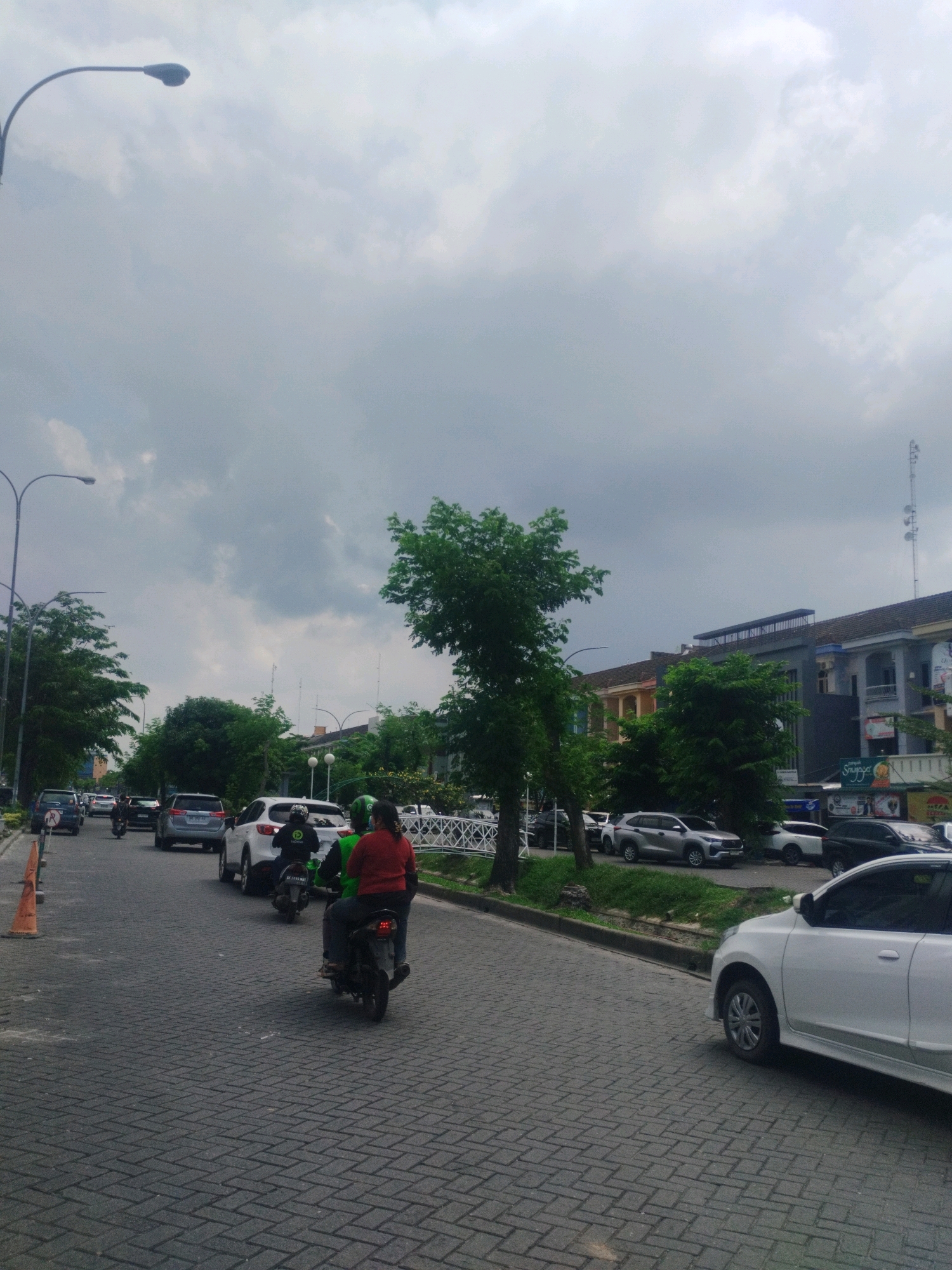 The image shows a street scene in Percut Sei Tuan, Indonesia, under a cloudy sky. The road is paved with dark interlocking bricks. Several cars are visible on the road, including a white car in the foreground on the right, a silver SUV, and other cars further back. Two people are riding a dark-colored motorcycle in the middle of the road, with the passenger wearing a red shirt and the driver wearing a green shirt. Trees line the right side of the road, and buildings with multiple stories are visible in the background. A lamppost is present on the left side of the image. Traffic appears to be moderate.