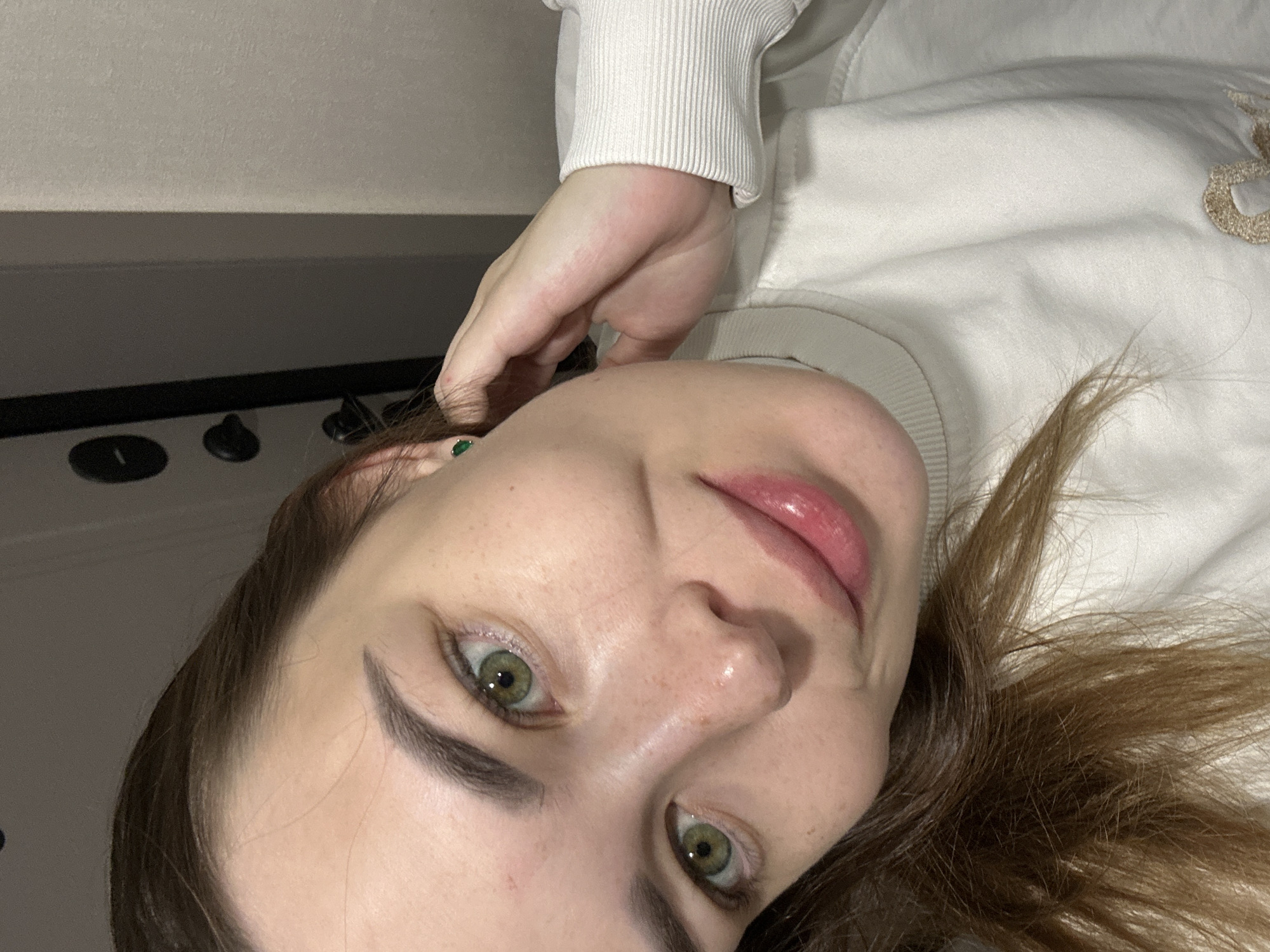 The image presents a close-up, tilted portrait of a young adult female, reportedly located in Perm, Russia. She has long, dark brown hair, parted on the viewer's right side, which falls partially over her shoulders. Her eyes are a prominent green, and her eyebrows are dark and well-defined. Numerous small, light brown freckles are visible across her nose and cheeks. Her lips are full, glossy, and tinted a soft pink, slightly parted in a gentle smile. She wears a cream-colored, ribbed turtleneck sweater, with a faint, light brown embroidered detail partially visible on the lower right chest area. A small, round, vibrant green stud earring is visible in her left earlobe (viewer's right). Her right hand (viewer's left) is raised, fingers curved, and rests behind her left ear, partially obscuring the left side of her face. The background, an indoor setting, features a dark, closed door with two circular knobs or locks on the left side of the frame, and a plain, light beige wall on the right.