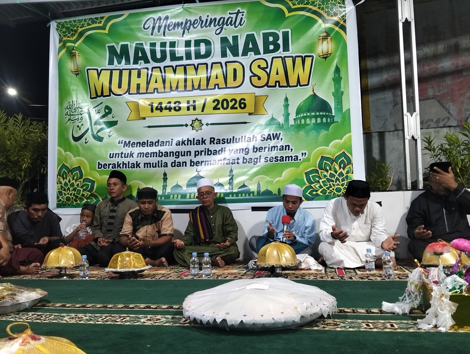The image depicts a gathering indoors, likely a community event. In the background, a large green banner with Islamic motifs and calligraphy is displayed. The banner reads "Memperingati Maulid Nabi Muhammad SAW 1448 H / 2026", indicating an observance of the Prophet Muhammad's birthday. Below the title, a quote in Indonesian translates to "Emulating the character of Rasulullah SAW, to build a faithful personality, noble character, and beneficial to fellow humans." Green ornate patterns and depictions of mosques and minarets are visible on the banner.
In the foreground, several men and a young boy are seated on a green carpeted floor, facing the banner. They are all wearing traditional Islamic attire, including peci (caps) and various styles of robes and shirts. Some of the men have their hands raised in prayer or supplication. In front of the seated individuals are several golden decorative domes and trays, some containing wrapped items, and bottles of water. The lighting suggests an evening or indoor setting. The overall atmosphere appears to be one of religious observance and community gathering.