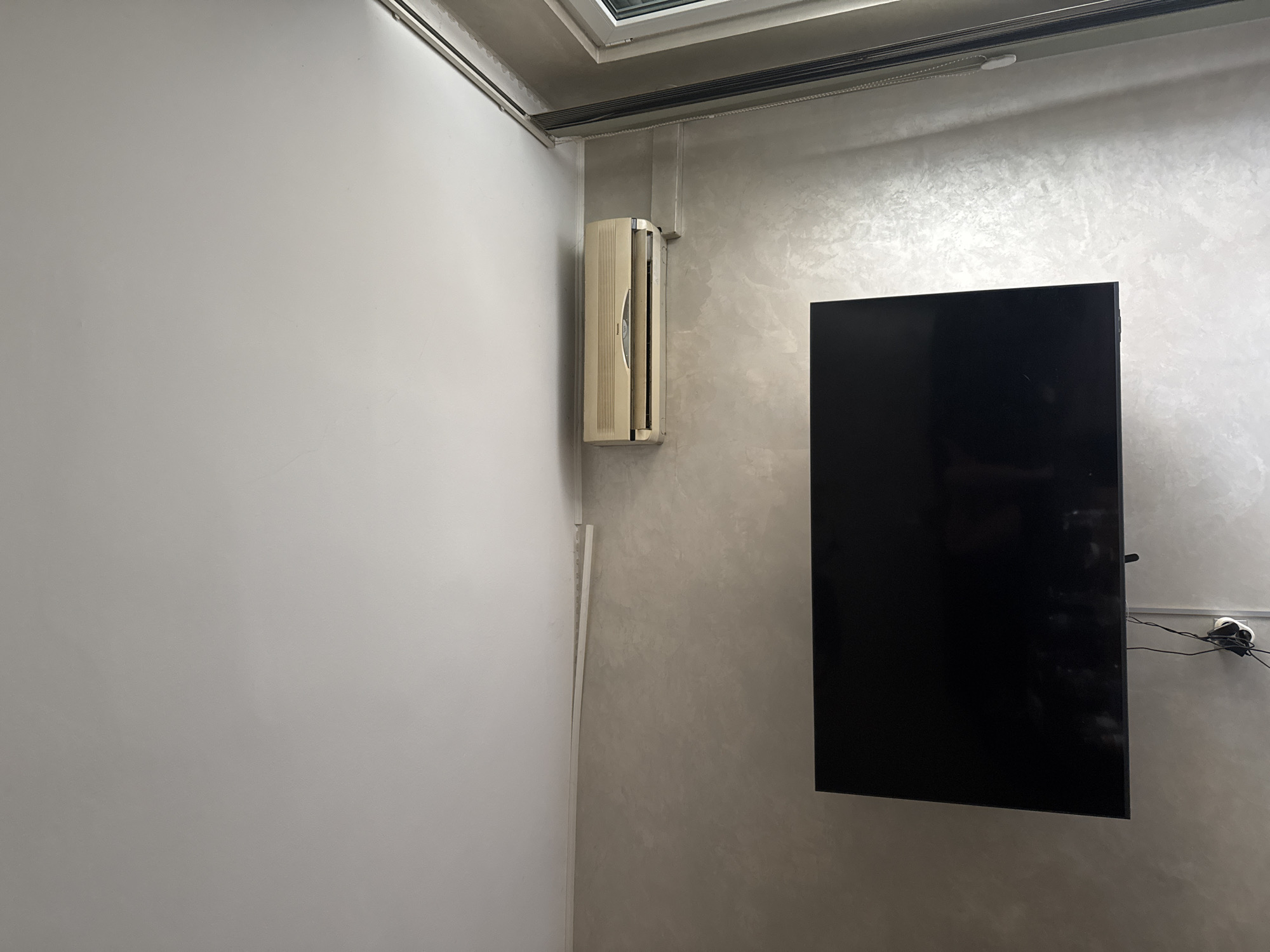 This indoor photograph captures a corner of a room, likely in Pleven, Bulgaria. The left wall is painted a plain white, contrasting with the right wall which features a light grey, textured finish, possibly stucco or Venetian plaster.
Mounted vertically on the textured wall, near the corner, is an older model off-white air conditioning unit. To the right of the air conditioner, a black flat-screen television is mounted on the same wall. Cables and wires are visible behind the television, connecting to a wall-mounted outlet. A white vertical conduit or trim piece runs down the corner where the two walls meet.
The ceiling above appears to be concrete, featuring a skylight or window that allows natural light into the space, indicating it is daytime. A set of blinds or a shade system is installed along the top edge of the textured wall, just below the ceiling, with a white bead chain visible for operation.
The scene is static, with no people or activities taking place. No discernible text is visible on any objects or surfaces. The overall impression is a functional, possibly modern or renovated, interior space.