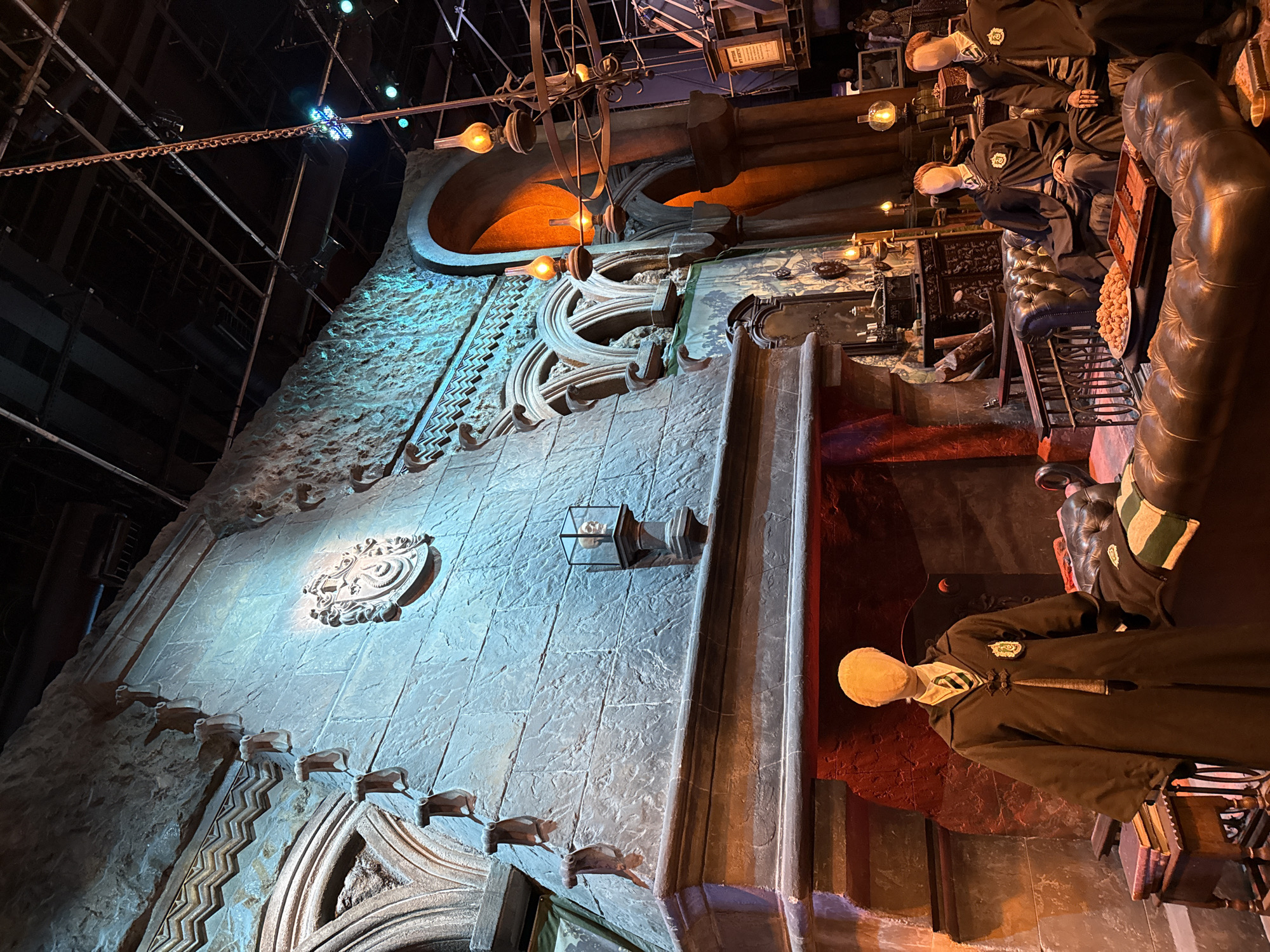 The image depicts a set resembling the Gryffindor common room from the Harry Potter films, located at Warner Bros. Studio Tour London – The Making of Harry Potter in Leavesden, near Watford, Hertfordshire, United Kingdom. The scene features a large stone fireplace, ornate archways, and a dark leather sofa. On the sofa, mannequins dressed in Gryffindor robes are seated. One mannequin stands by the fireplace, also in a Gryffindor robe. A coffee table in front of the sofa holds a plate of pastries and a game board. Books are stacked on a side table near the fireplace. The lighting is warm and atmospheric, with lamps casting an orange glow. The background shows more of the studio set's construction and lighting rig. The overall impression is of a meticulously recreated cinematic environment.