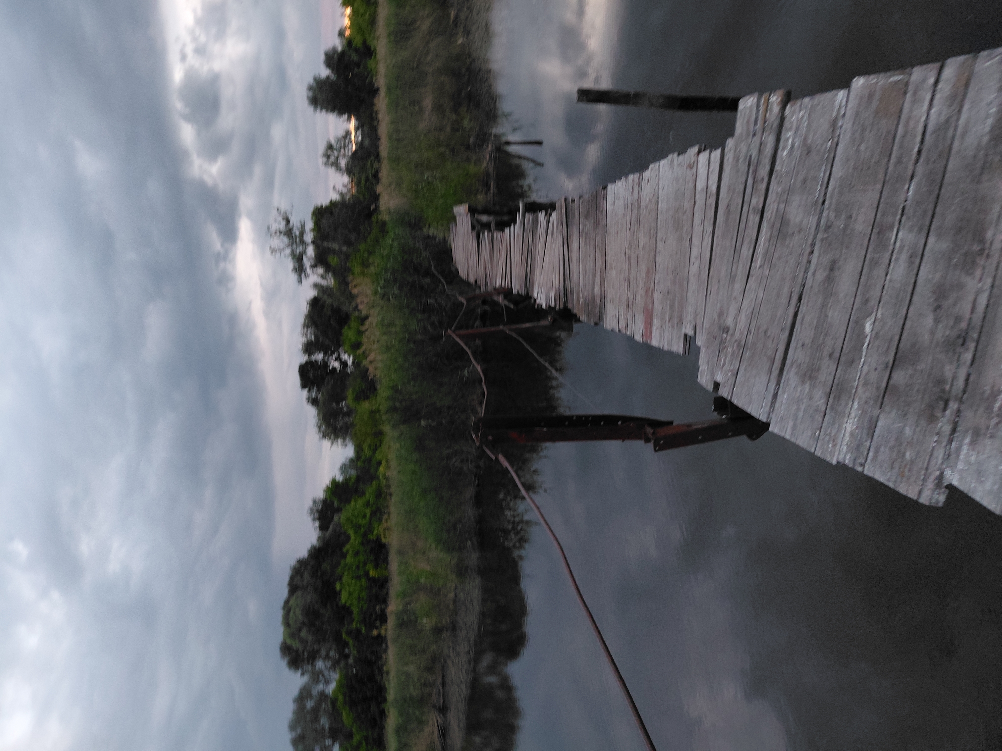 A weathered wooden footbridge extends across dark, still water towards a line of trees under a cloudy sky. The footbridge, constructed of aged planks, is angled from the lower right to the upper left of the frame, appearing somewhat dilapidated. A rusty wire railing runs along the left side of the bridge, supported by wooden posts. The water reflects the overcast sky and the surrounding reeds and trees. The environment appears to be a natural setting, possibly a marsh or riverbank, in Ustyvytsia, Ukraine. No people are visible in the scene. The overall mood is somber, due to the dark, cloudy conditions.
