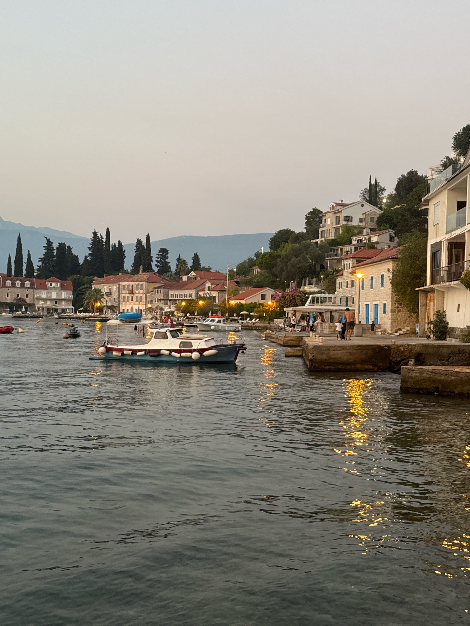 A body of water with a small boat in the foreground is shown. The boat is white with a red stripe and has buoys attached to its sides. In the background, there are buildings lining the shore, some of which are hillsides. The buildings are of various colors and architectural styles. Trees and mountains are also visible in the distance. The sky is overcast and the lighting suggests it is either dawn or dusk. The water reflects the lights from the buildings, creating streaks of yellow and orange. There are several people on the shore near the buildings.