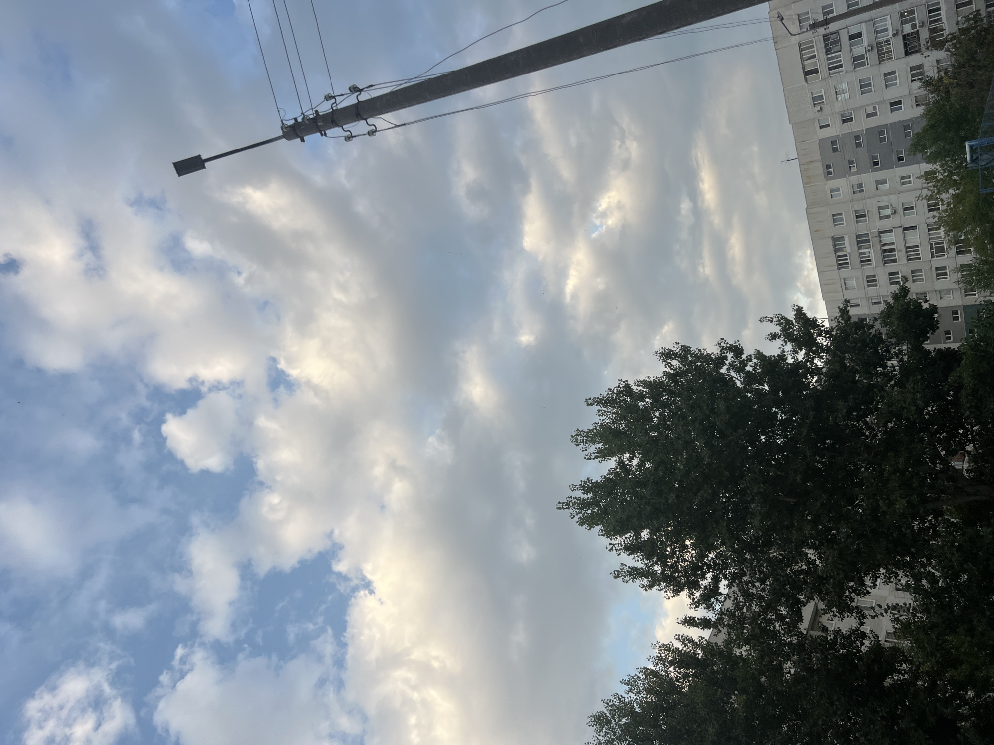 The image depicts a cloudy sky above a residential building and trees in Ulyanovsk, Russia. A tall, concrete utility pole with electrical wires and a street lamp stands on the right side of the frame. In the lower portion of the image, dark green trees are visible in front of a multi-story apartment building. The building appears to be constructed of light-colored concrete and features numerous windows. The sky is filled with a mix of white and grey clouds, with patches of blue visible.