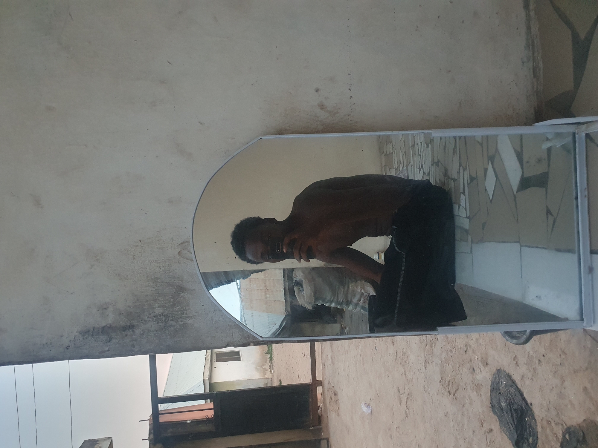 A young man is captured in a mirror selfie, shirtless or wearing a very dark top and dark trousers, holding a smartphone to take the picture. The mirror is a tall, white-framed freestanding type.
The scene appears to be an outdoor or semi-outdoor space in Lokoja, Nigeria, likely during the daytime under clear weather. The background reflected in the mirror behind the man shows a light-colored wall and some indistinct objects, possibly white sacks.
Outside the mirror's reflection, the immediate environment features a textured, light-colored wall on the left and top, possibly concrete or painted plaster, showing some signs of wear or discoloration. The ground to the right and below the mirror is a patterned surface of light-colored tiles or paving stones, and a dark, crumpled object lies on the ground near the base of the mirror. In the lower left part of the image, beyond the mirror, an outdoor view reveals what seems to be other buildings under a bright sky and some metal railings or structures. The rough textures and outdoor elements are consistent with a utilitarian or residential exterior setting. No visible text is present in the image.