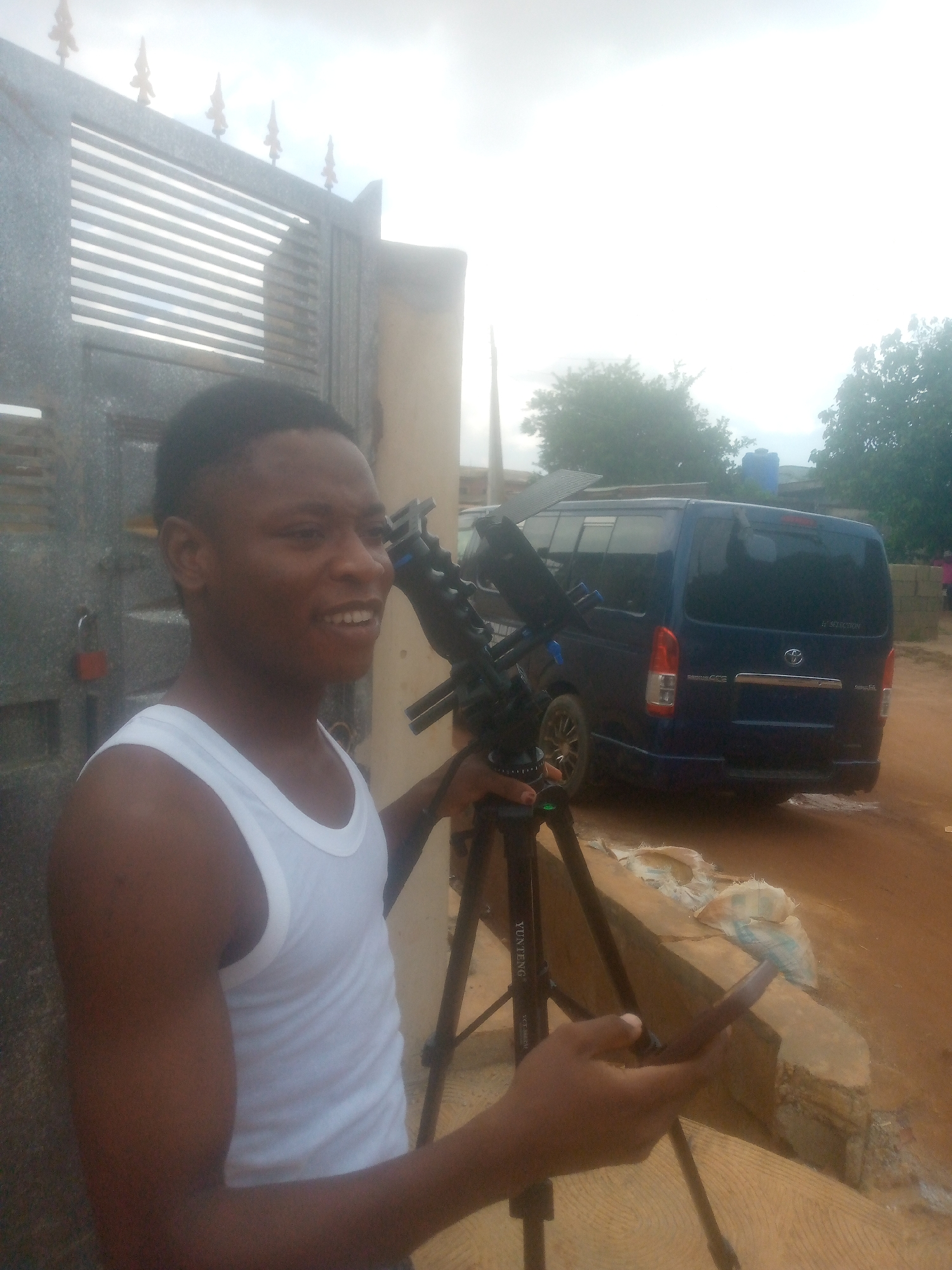 The image shows a man standing outdoors next to a gate, holding a phone in his left hand and a tripod on his right. He is wearing a white tank top and has short, dark hair. Behind him is a dark blue Toyota van parked on a dirt road. The location appears to be a residential area, possibly a suburban or rural setting, given the dirt road and the style of the surrounding buildings. The sky is overcast.