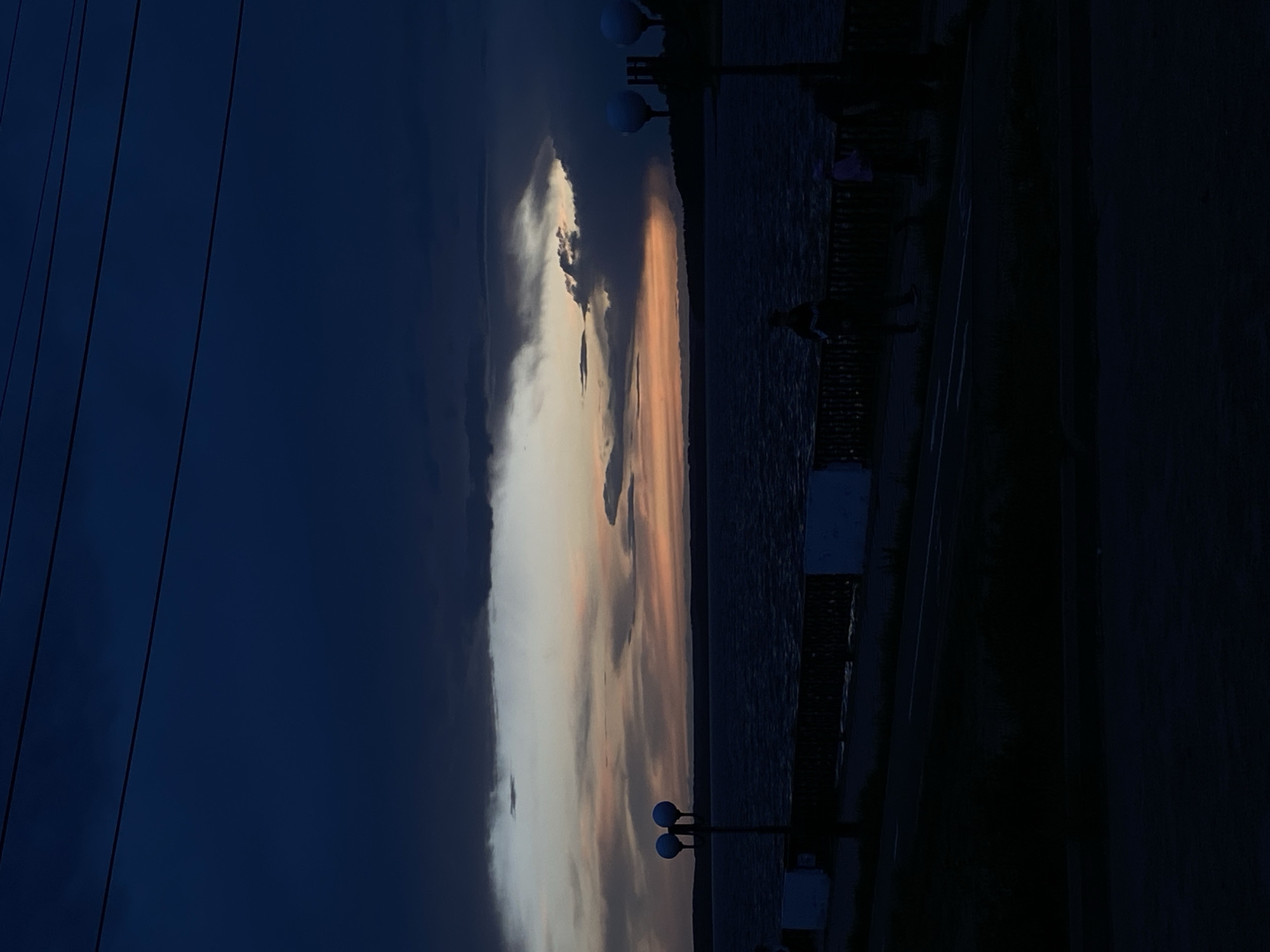 The image depicts a scene at dusk or dawn in Votkinsk, Russia, possibly along the Votkinsk Reservoir. The sky exhibits a gradient of colors, ranging from deep blue on the left to lighter shades of white and orange near the center where the sun appears to be setting or rising. The presence of clouds adds texture and complexity to the sky.
A body of water, presumably the Votkinsk Reservoir, occupies the central portion of the image. The water surface appears relatively calm, reflecting some of the sky's colors.
Along the right side of the frame is a promenade or walkway, indicated by the presence of lampposts and what appears to be a low railing or fence. Silhouetted figures of people can be seen walking along the promenade.
Power lines are visible on the left side of the image.