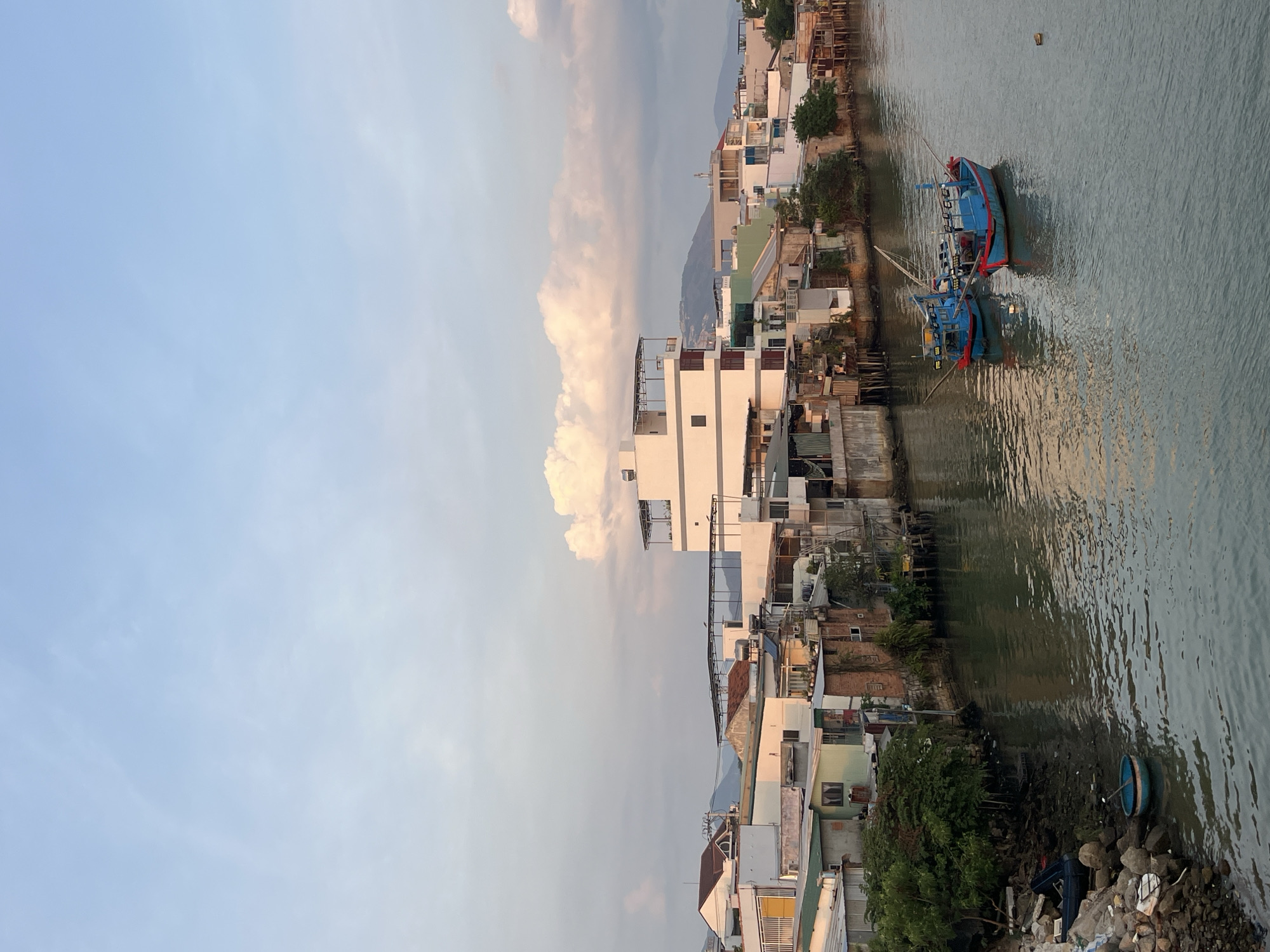The image captures a waterfront scene in Bac Nha Trang Ward, Vietnam. Predominantly, the frame displays a collection of buildings, varying in height and architecture, situated along the water's edge. A pair of blue and red fishing boats are moored along the waterfront. The water itself is calm. The sky is partly cloudy, with some large cumulonimbus clouds, and the lighting suggests either early morning or late afternoon. The overall impression is that of a bustling waterfront community.