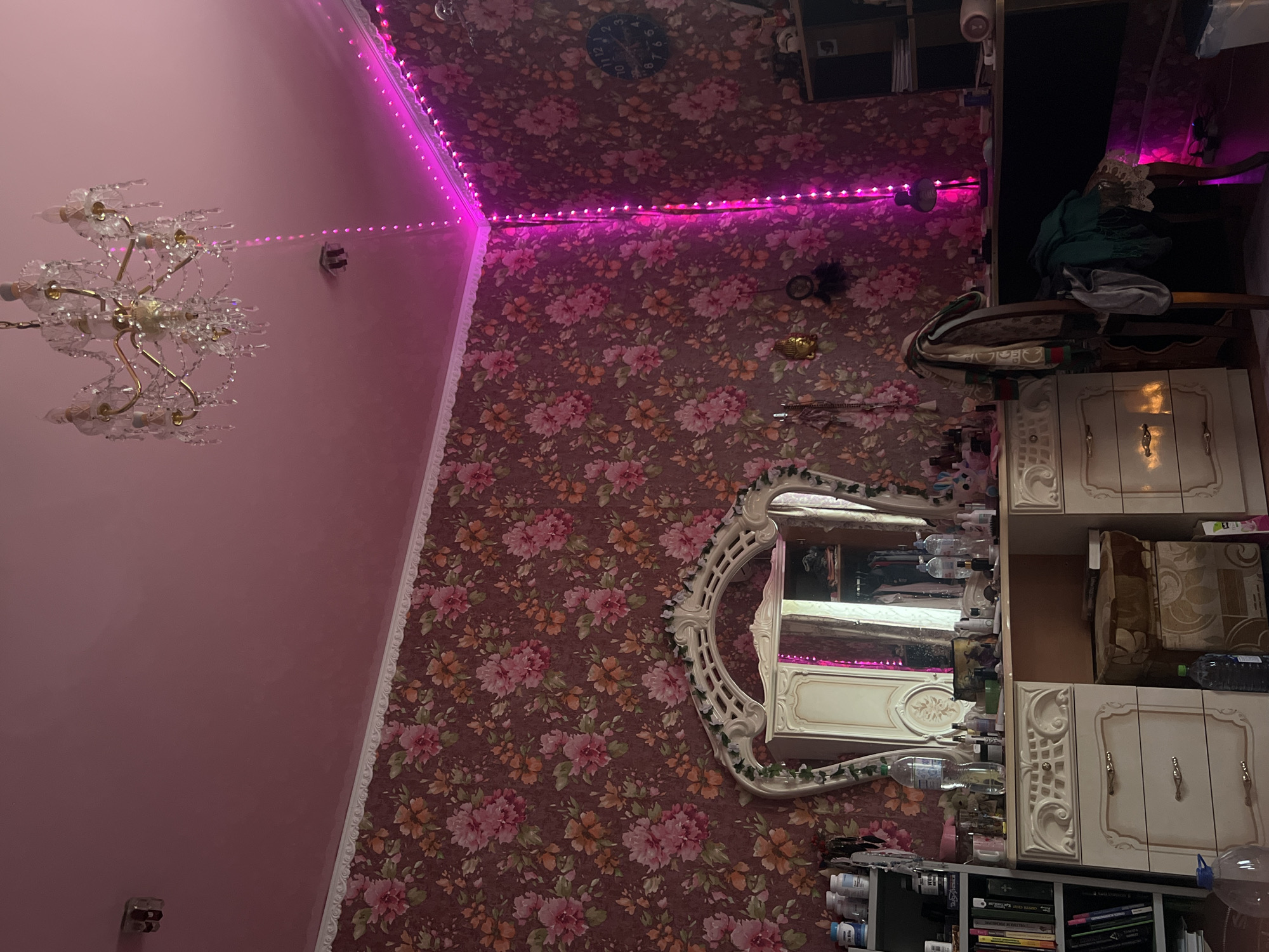 The image depicts a dimly lit bedroom with pink-toned walls and floral wallpaper. A large, ornate vanity with a mirror dominates the left side of the frame, featuring drawers and various bottles and containers. To the left of the vanity, a bookshelf is filled with books and other items. Above the vanity, a crystal chandelier hangs from the ceiling. Pink LED strip lights are installed along the ceiling edges in the corner of the room, casting a vibrant glow.
On the right side of the image, a dark desk is visible, cluttered with items. A chair is placed at the desk, draped with clothing. Further to the right, a shelving unit is partially visible, containing more items. A wall clock with a dark blue face is mounted on the wall, displaying the time as approximately 10:10. The overall impression is of a decorated, somewhat cluttered personal space.
This scene is located in the city of Krasnoe-na-Volge, Russia.