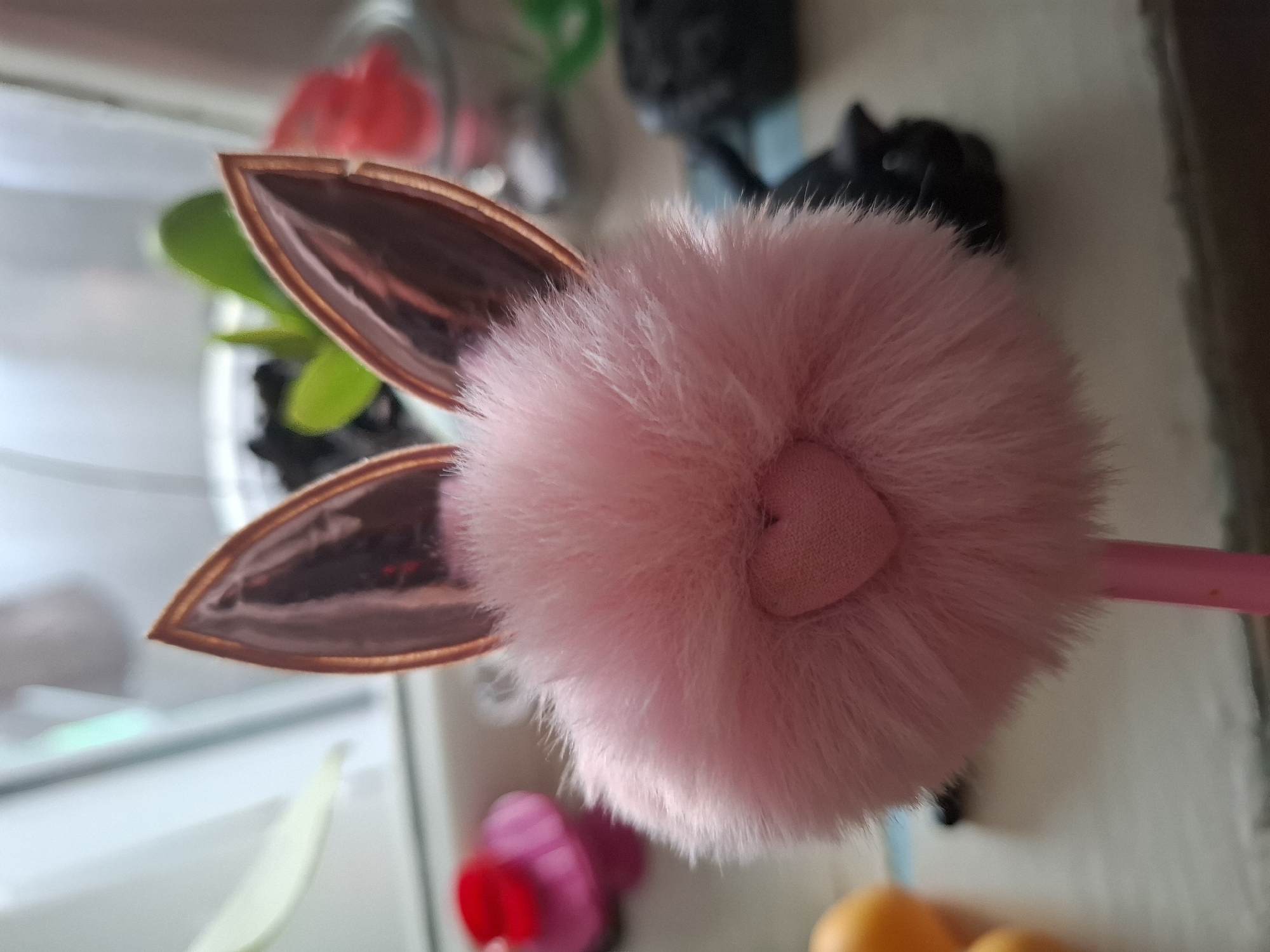 The image shows a close-up of a pink, fluffy pom-pom attached to two rose-gold colored bunny ears. The pom-pom has a small heart-shaped detail in its center. The bunny ears are pointed and metallic, reflecting light. In the background, there are out-of-focus elements suggesting an indoor setting. These include a small green plant in a clear container, some red and pink objects, and what appears to be a black figurine. The lighting suggests it is daytime, possibly indoors near a window due to the reflections and soft light. The overall mood is playful and whimsical, likely due to the animal-themed accessory.
