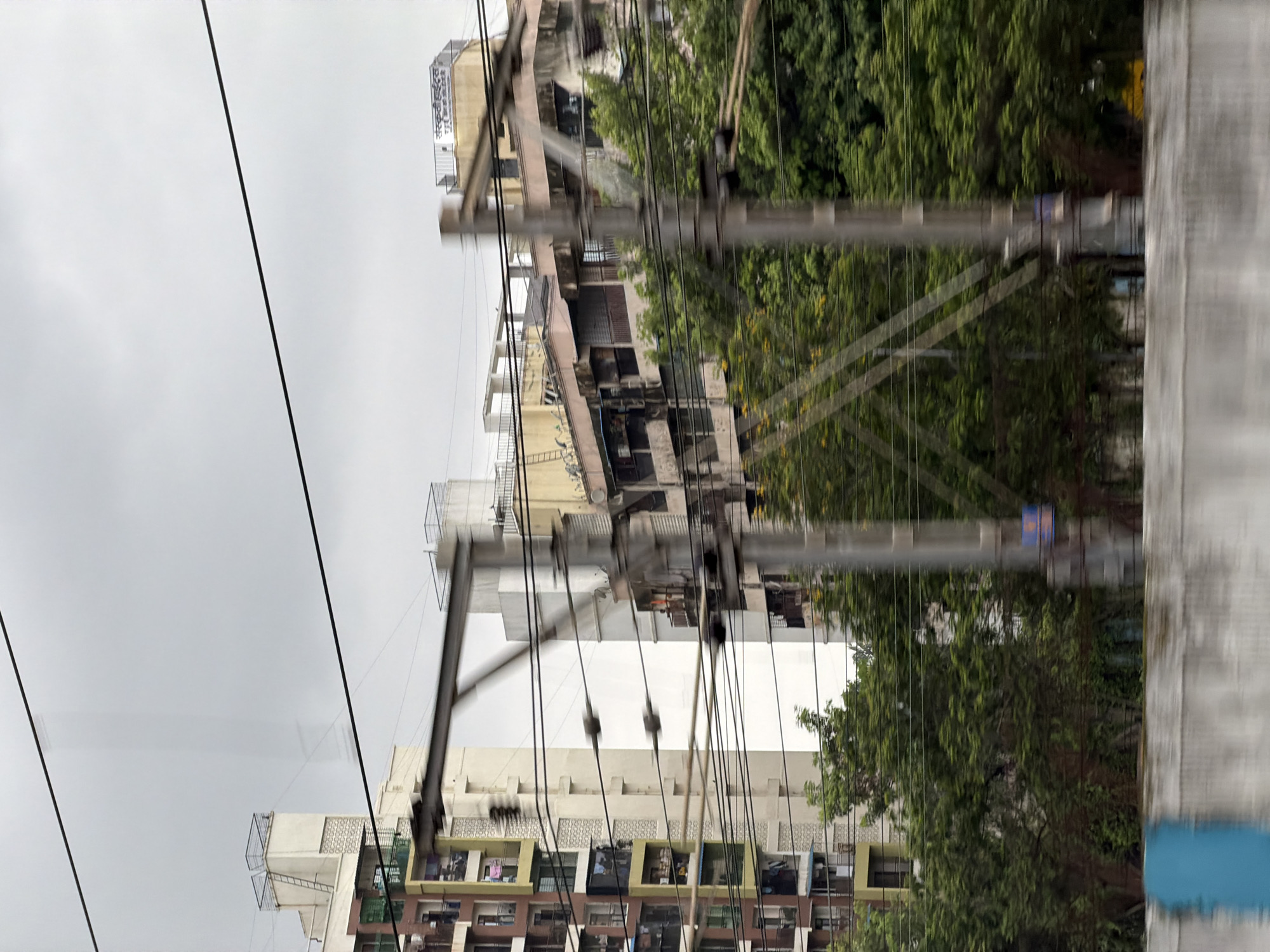 The image shows a view of buildings and trees in the city of Vasai-Virar, India. The buildings are multi-story apartment complexes, some of which appear to be under construction or in disrepair. There are many electrical wires and utility poles crisscrossing the scene, suggesting an urban environment. Lush green trees are visible in the foreground and interspersed between the buildings, indicating a good amount of vegetation. The sky is overcast with gray clouds. The image appears to have been taken from a moving vehicle due to the motion blur. There are no people or any visible actions or interactions occurring in the scene.
