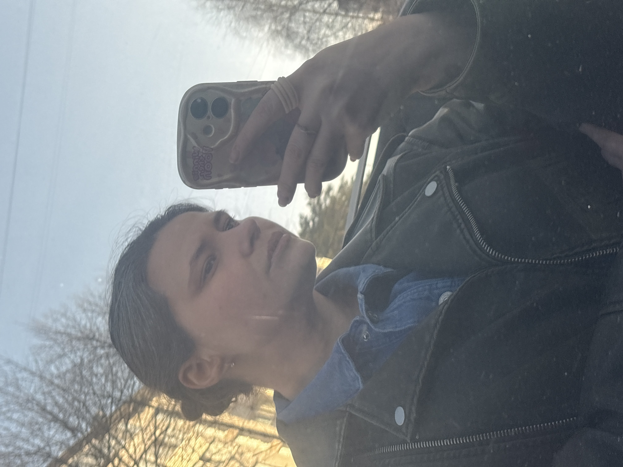 A person is taking a selfie. The person is wearing a black leather jacket over a blue denim shirt and is holding a phone with a phone case in their left hand. The background is slightly blurred but appears to be trees and buildings. The location is plausibly in a city, potentially Chaikovsky, Russia, given the urban elements and foliage.