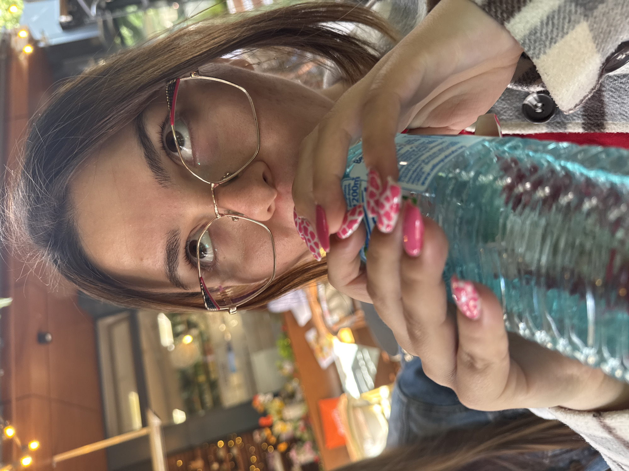 A person with brown hair and eyeglasses holds a water bottle with pink and white patterned nails. The person is wearing a plaid jacket over a red shirt. The location appears to be an outdoor seating area in an urban setting, with blurred background details of buildings, plants, and lighting. The city is Unknown, Unknown.
