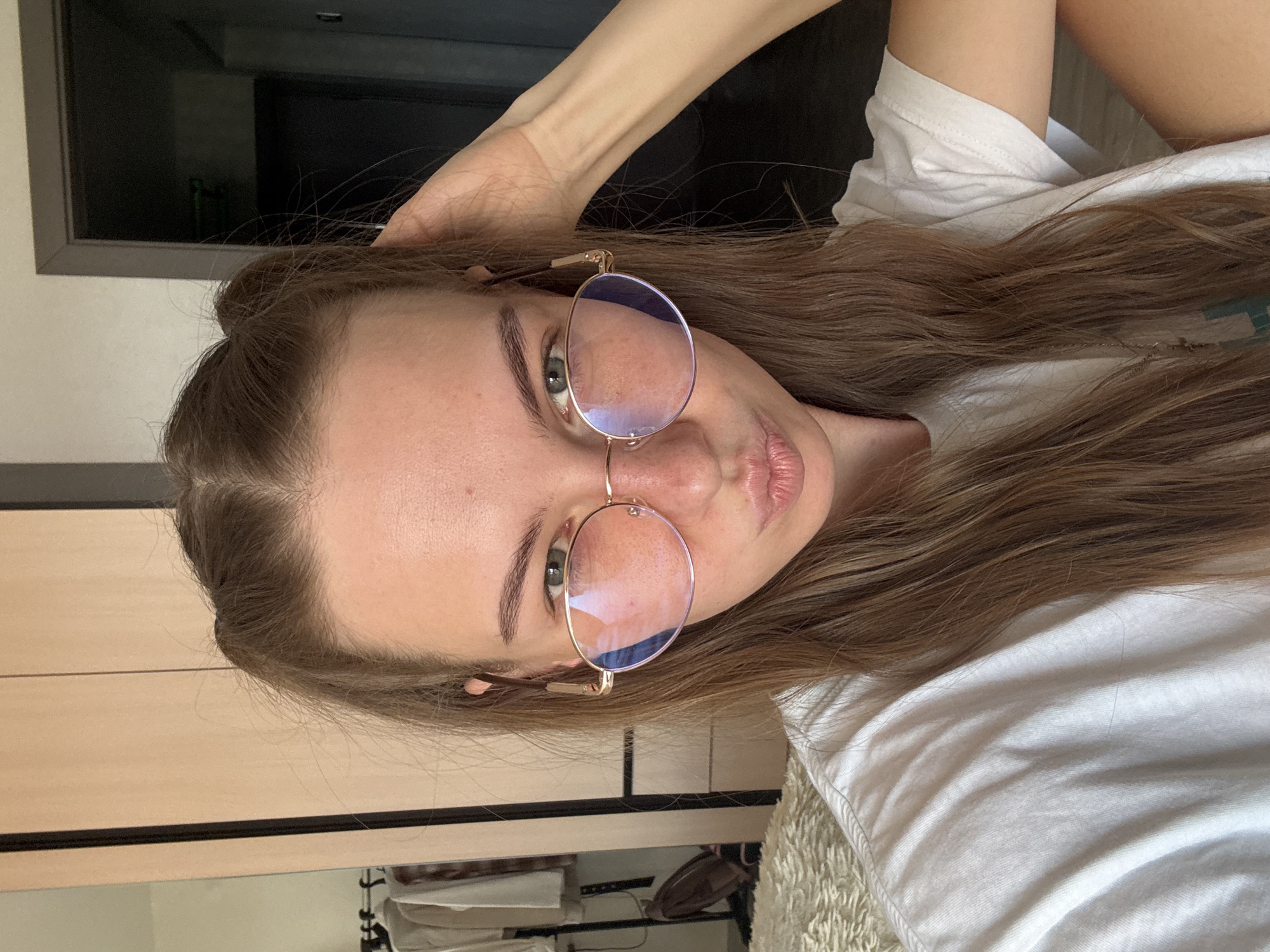 A young woman with long, light brown hair, styled in two pigtails, is looking at the camera and puckering her lips. She is wearing round, gold-rimmed glasses and a white t-shirt. Her right hand is resting on the back of her head. The background shows a closet with light wood doors and a slightly blurred reflection in a mirror to the left. There is also a doorway visible in the background to the right. The setting appears to be a bedroom.