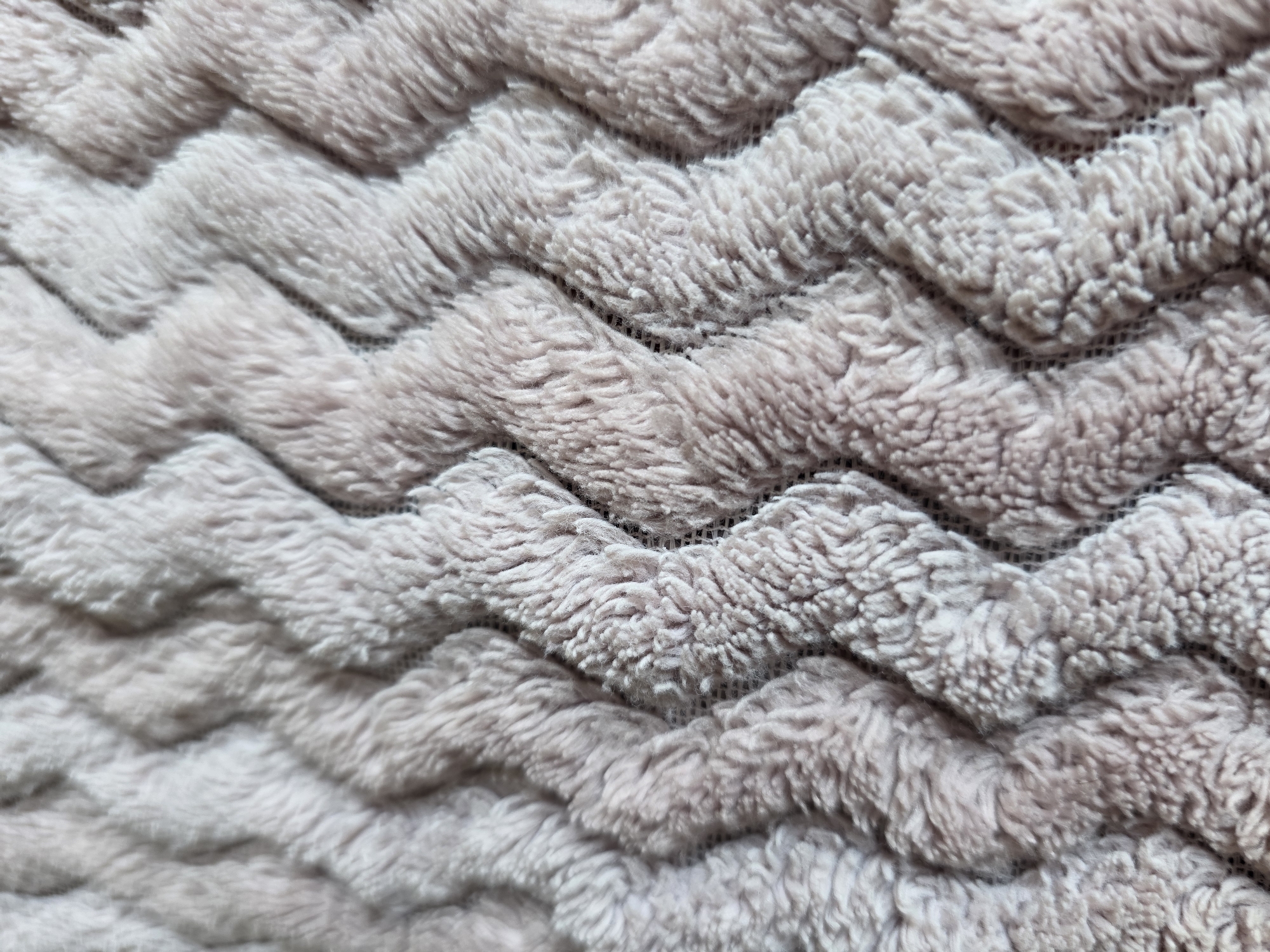 This media file is a close-up, abstract shot of a textured, fuzzy fabric with a chevron pattern. The fabric appears to be a soft, plush material, possibly a blanket or upholstery. The color is a muted, dusty rose or light mauve. The chevron pattern is created by raised ridges of the fabric, with darker, recessed areas between them, suggesting a woven or knitted construction. The lighting highlights the texture, casting subtle shadows that emphasize the raised elements. There are no people, objects, or a discernible scene or setting. No activity or event is taking place. The image is focused entirely on the textile's surface. No text is visible. The location context of Kyiv, Ukraine, does not provide any specific visual cues within the image itself.