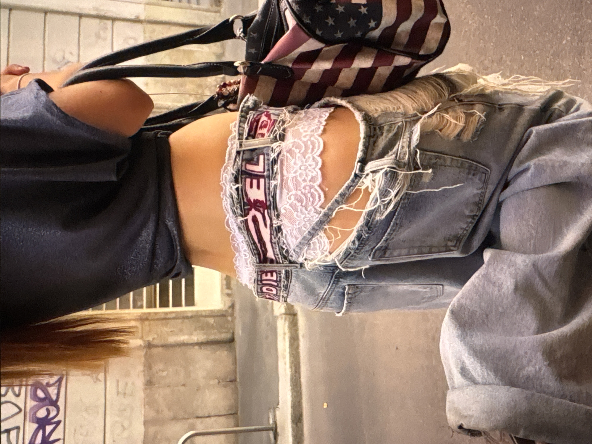 A person is captured from the waist down, wearing distressed denim jeans and a dark, loose-fitting top. The jeans have significant rips and tears, exposing lace-trimmed underwear. A handbag with an American flag pattern rests on the person's hip. The scene appears to be outdoors, possibly on a street, with a wall and some graffiti visible in the background. The lighting suggests it could be daytime. The person's posture and the overall styling indicate a casual, perhaps edgy, fashion statement. The visible text on the underwear band reads "DIE".