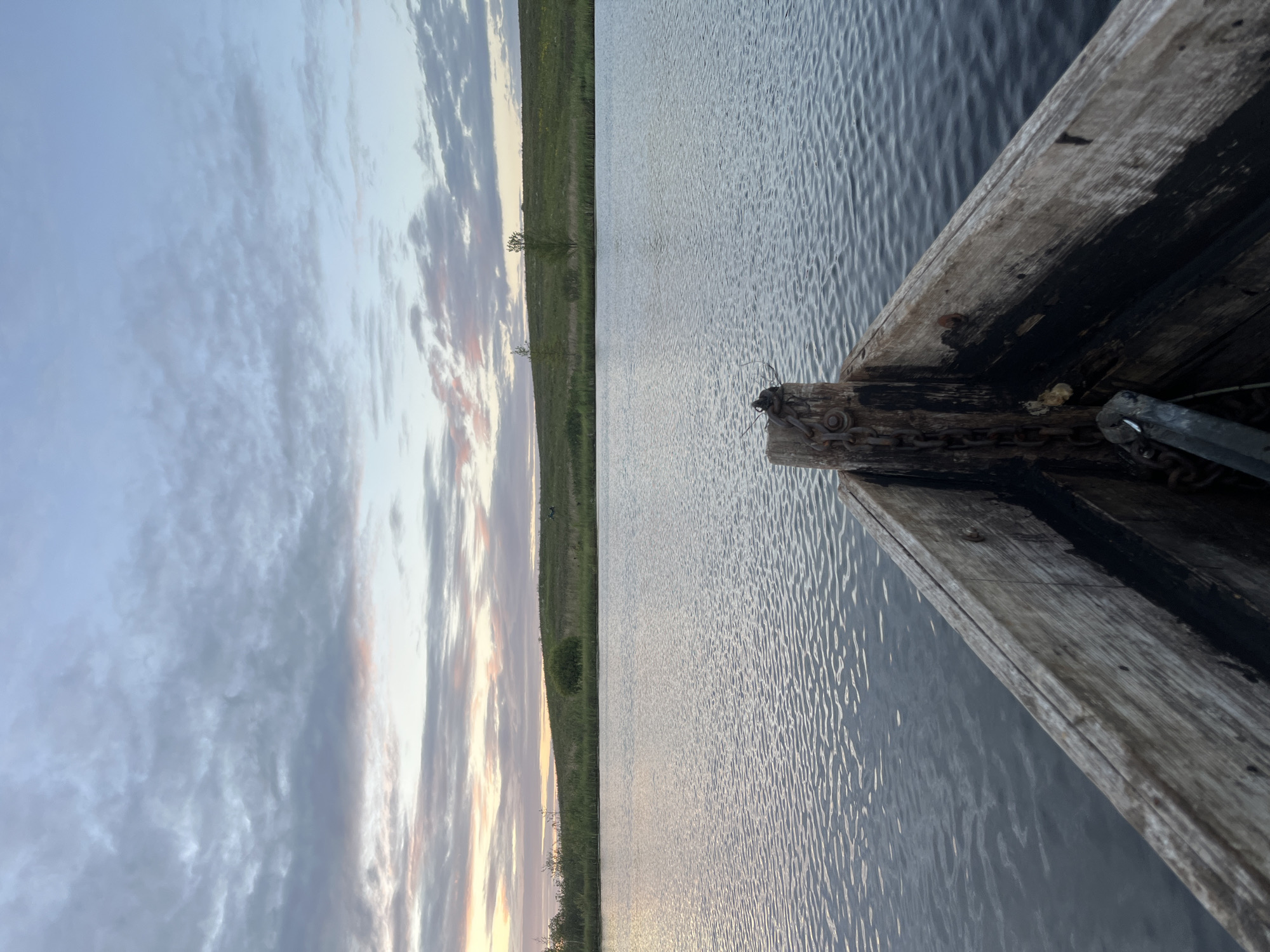 A view from the front of a weathered wooden boat on a calm body of water suggests a serene natural setting. The water's surface is gently rippled, reflecting the soft hues of a cloudy sky. The sky itself transitions from a pale blue at the top to warmer pink and orange tones near the horizon, indicating either sunrise or sunset.
Beyond the water, a grassy embankment rises, dotted with sparse trees and bushes. The landscape appears rural and undeveloped, consistent with a location like Дунілавіцкі сельскі Савет, Belarus. The focus is on the natural elements – the sky, water, and land – with the boat providing a foreground perspective. There are no people or animals visible in the frame, and no specific actions are depicted beyond the implied stillness of a boat at rest. The overall atmosphere is one of quietude and natural beauty.
