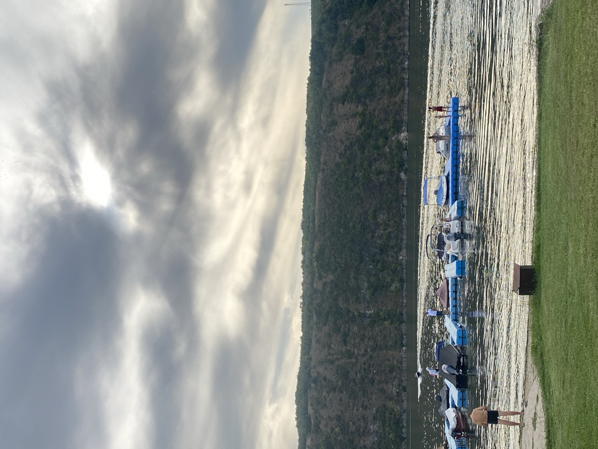 The image depicts a waterfront scene in Molovata Nouă, Moldova, featuring a lake or river with a steep, tree-covered hill in the background. A floating dock system is positioned on the water, equipped with several covered boats and a jet ski. A jet ski is visible in motion on the water in the distance.
On the dock, there are multiple individuals. Two people are standing on the dock, and another individual is visible on the dock further in the background. Closer to the shore, on the dock, a group of three individuals are present.
In the foreground, a shirtless man wearing black shorts stands on a grassy area near the water's edge, facing away from the camera. A dark brown box-like object is situated on the grass to his right. The water exhibits rippling patterns reflecting the cloudy sky. The sky itself is overcast with dramatic clouds, and the sun is partially obscured, creating shafts of light. The vegetation on the distant hill appears dense and varied in color, with some patches of dry grass or soil visible. A tall tower structure is faintly visible on the crest of the hill on the right side of the frame.