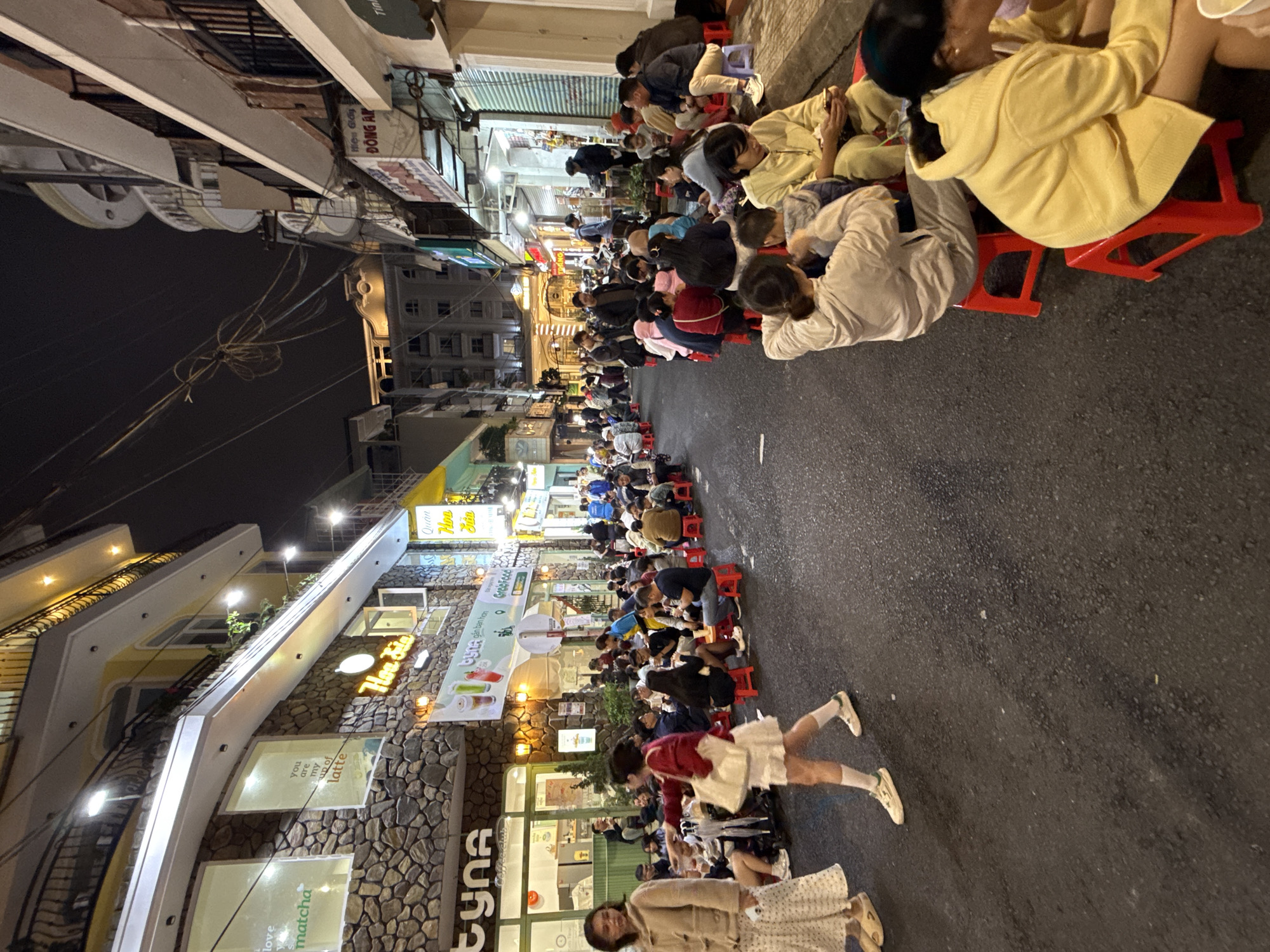 This image captures a vibrant night scene on a bustling street in Da Lat, Vietnam. The setting is an urban environment, likely a food or pedestrian street, lined with multi-story buildings housing various shops and cafes.
The main subjects are numerous people, predominantly young adults, who are gathered on the asphalt street. They are seated closely together on small, low red plastic stools, a common sight in Vietnamese street food culture. Many individuals are engaged in conversation, looking at their phones, or enjoying food and drinks. The atmosphere appears lively, social, and relaxed, indicative of a popular gathering spot. Two young women are prominent in the foreground, one in a red jacket and white skirt seeming to interact with someone off-camera, and another in a light-colored dress and coat looking towards the camera.
Notable details include the time of day, which is clearly night, illuminated by numerous streetlights and the bright signs of establishments. The weather appears clear. The street is busy, with rows of people extending into the distance, suggesting a well-attended event or popular evening spot. The architecture includes stone-clad facades and modern building designs. Overhead, power lines and street lighting are visible.
Visible text on storefronts and signs includes "TYNA," "Quán Hoa Jin," "Hiệu Giấy ĐÔNG AN," and phrases like "love matcha" and "you are my cup of latte," indicating cafes or beverage shops. There's also "Chi gần bạn hơn" and "Tin." The scene perfectly encapsulates the energetic street life characteristic of many Vietnamese cities, offering a glimpse into local social customs.