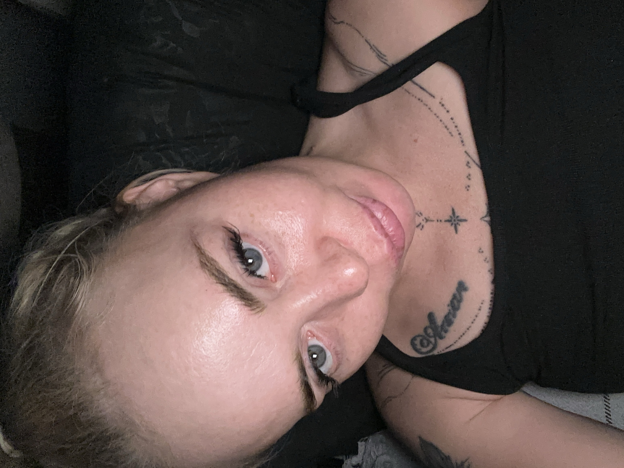 A close-up, high-angle, indoor selfie shows a young, Caucasian woman with blonde hair pulled back, and striking blue-grey eyes. She has fair skin with visible pores and some light freckles. Her eyelashes are enhanced with dramatic false lashes. Her lips are full and appear slightly parted. She is wearing a black tank top, revealing tattoos on her chest and shoulder. The tattoo on her chest reads "Amman" in cursive script, above a dotted line and a star-like design. Another tattoo with lines and dots is visible on her right shoulder. The background is dark and appears to be a soft, textured surface, possibly a chair or blanket. The lighting is dim and creates soft shadows on her face. The setting is indoors in an unidentifiable location.