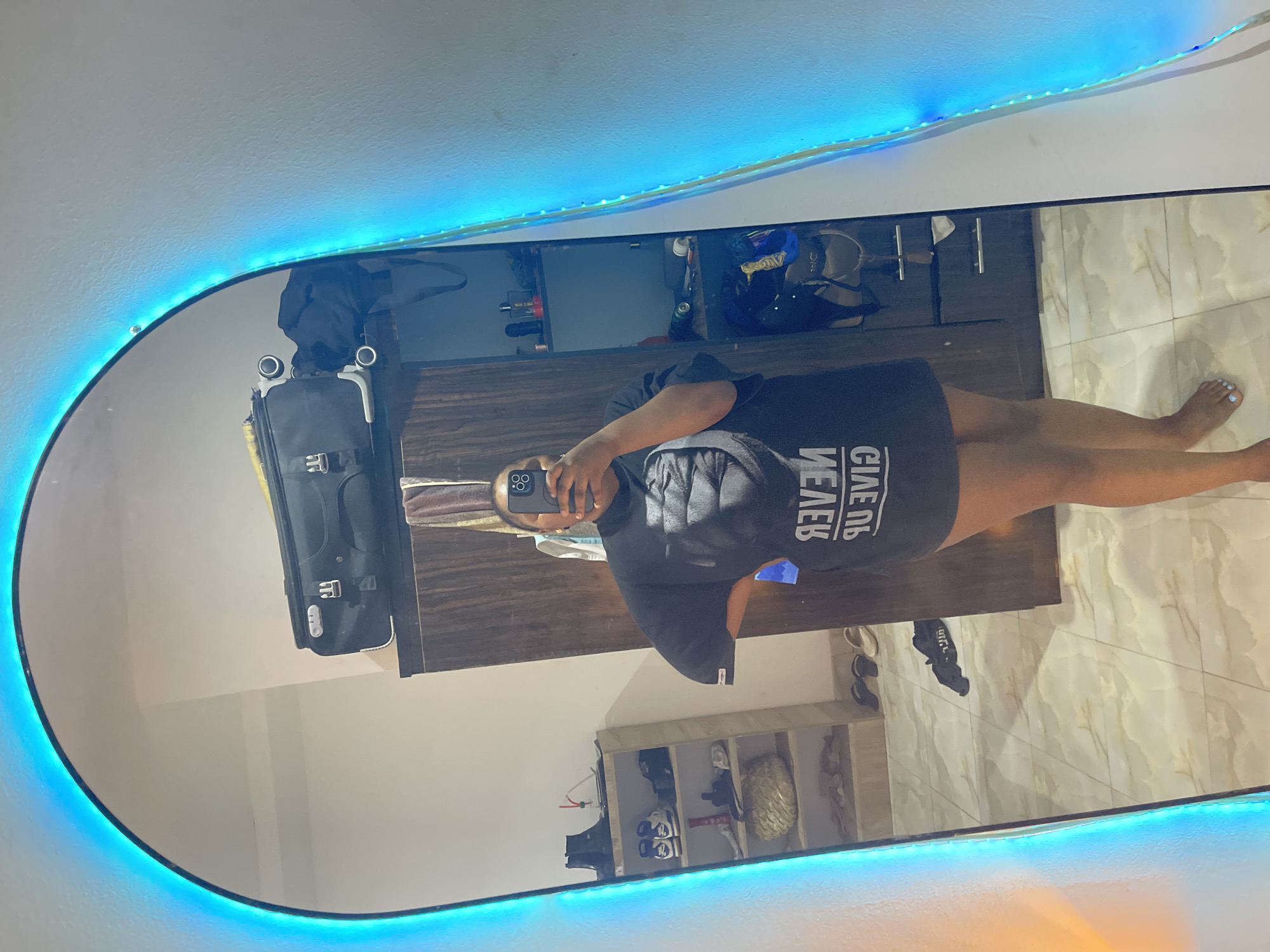 A person, seen in a mirror selfie, stands in what appears to be a bedroom or closet area. The person is holding a smartphone and is partially visible in the reflection, wearing a dark t-shirt. A luggage bag is leaning against the wall next to a wooden cabinet. A display shelf unit with shoes and miscellaneous objects is visible behind the person. The floor is tiled in a light color. The mirror is illuminated with a strip of blue LED lights. It's not possible to discern the location in Abuja, Nigeria, from this image alone.