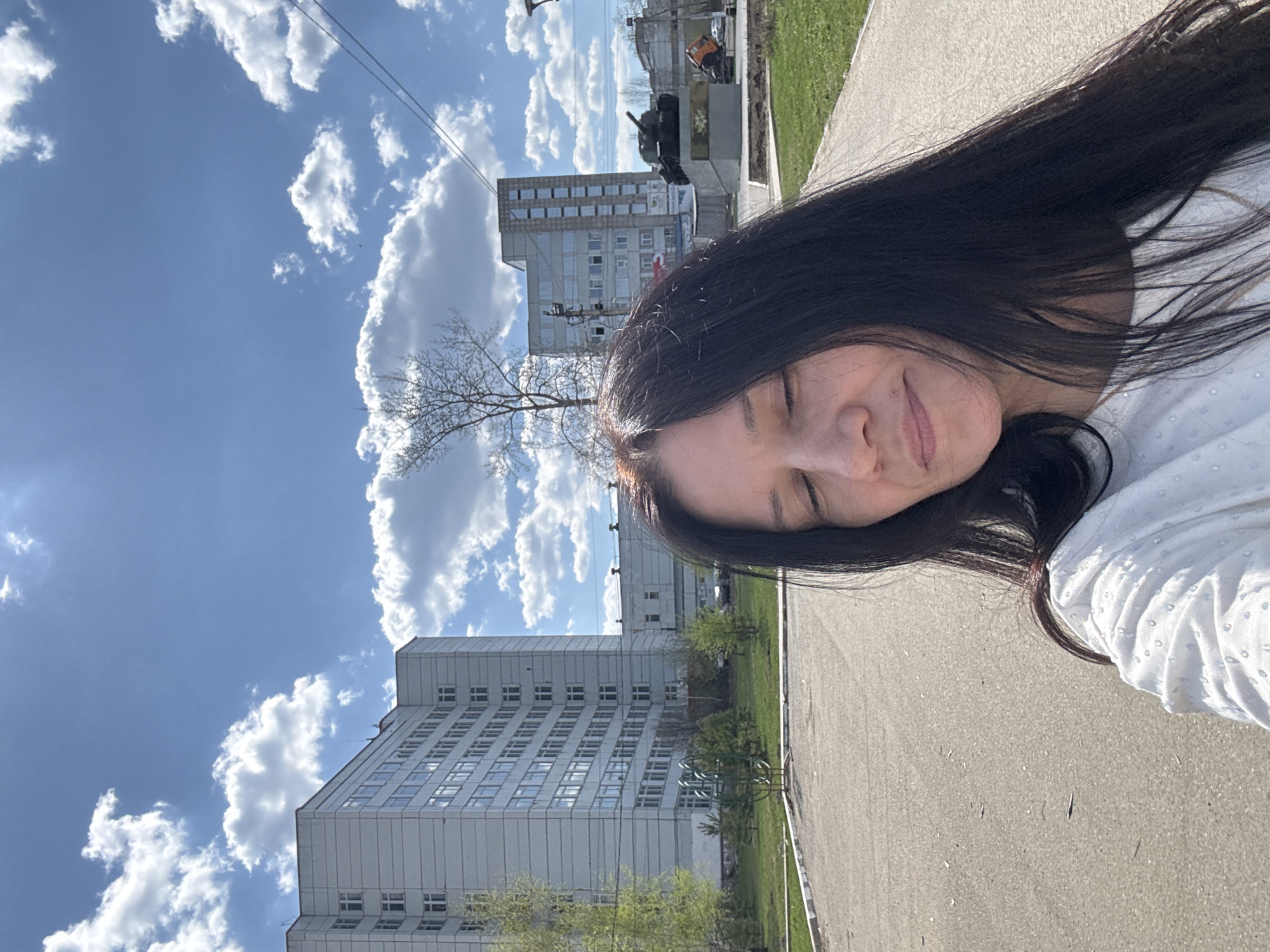 A selfie of a woman with long, dark hair is the central subject of the image. She is wearing a white blouse with a textured pattern. The background shows an urban scene, including a building with numerous windows, indicating a residential or commercial structure. A street or sidewalk runs alongside the building. In the distance, there is a war memorial, suggesting the location is likely in a public space. Fluffy white clouds are visible in the blue sky. It appears that the photo was taken outdoors on a sunny day, and the location is in the city of Chaikovsky, Russia.