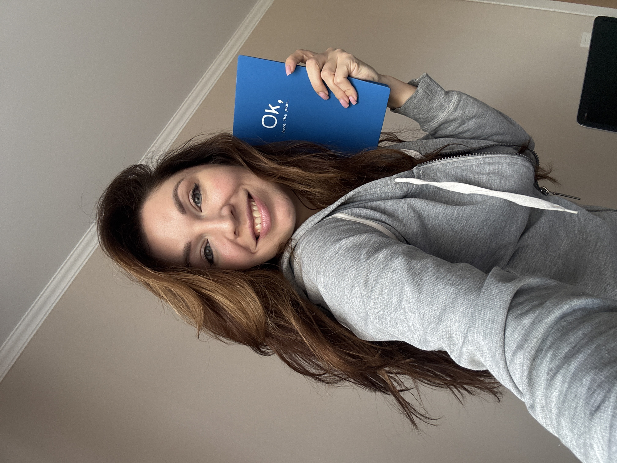 A woman with long brown hair and a gray zip-up hoodie smiles while holding a blue book with the words "Ok, here's the plan..." written on it. She is indoors, presumably in a room with beige walls and white crown molding near the ceiling. A black electronic device is visible to the upper right of the frame.