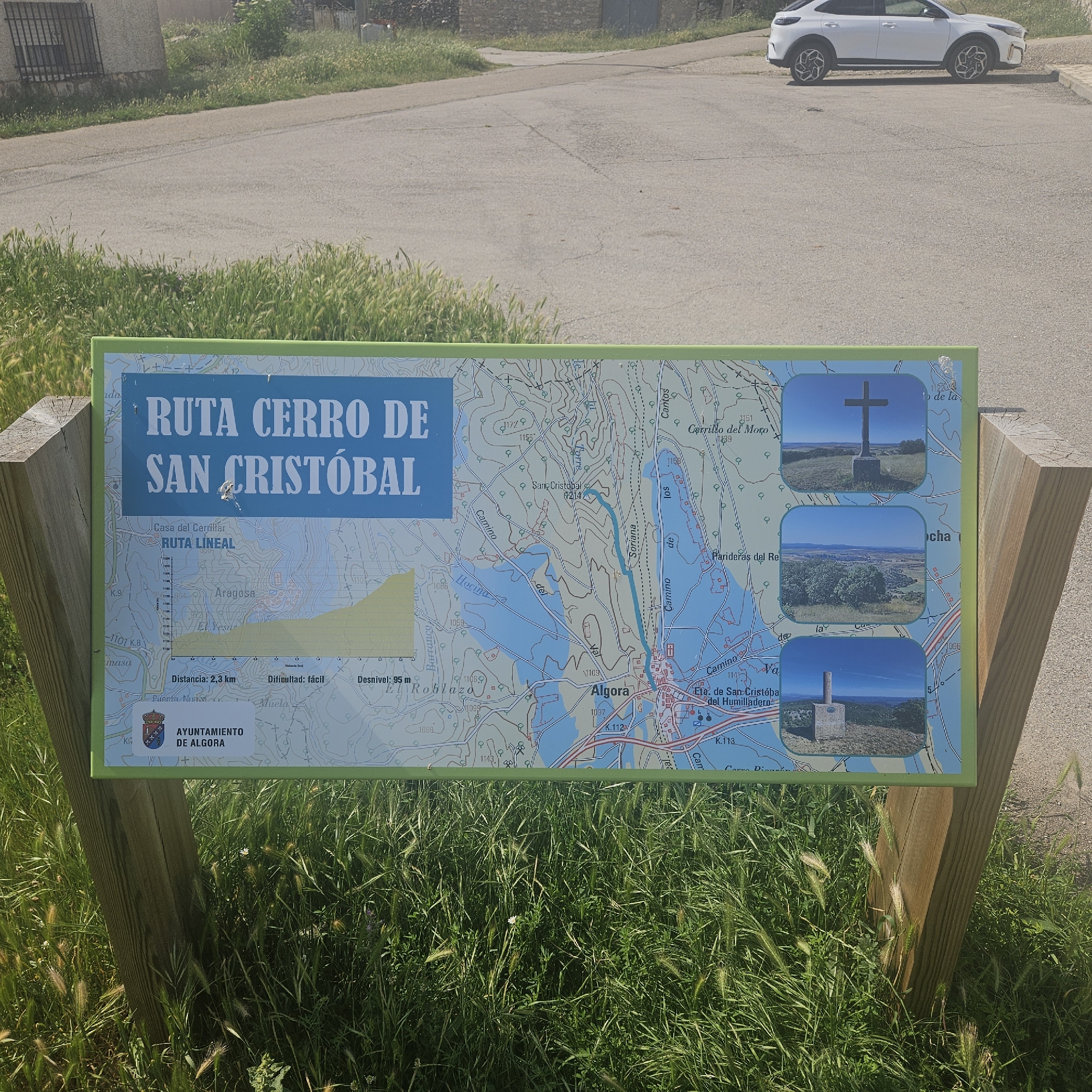 The image shows a sign indicating the "Ruta Cerro de San Cristobal" (San Cristobal Hill Route), located in Algora, Spain. The sign features a map of the linear route, with a blue path illustrating the trail, along with the location of San Cristobal at the top of the hill. The map shows the general area of Algora as well. The sign also provides additional information such as the distance of the route (2.3 km), the difficulty level (easy), and the elevation gain (95 m). Additionally, the sign features photographs of landmarks along the route, including the summit cross. The coat-of-arms of the City of Algora appears on the lower left. The sign is supported by wooden posts, and surrounding the base of the sign is vegetation. In the background, a white vehicle is parked on an asphalt surface near a building.
