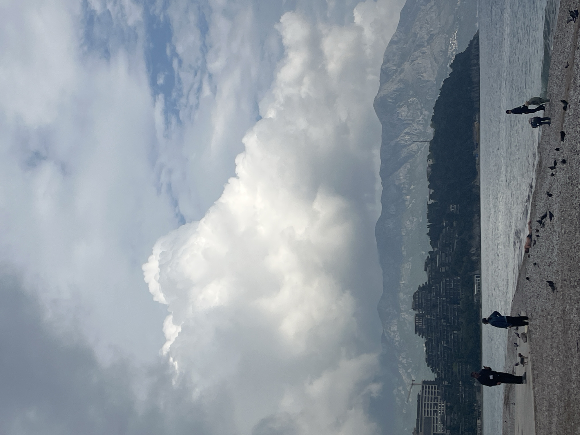 A panoramic coastal scene in Budva, Montenegro, unfolds under a dramatic sky. The upper portion of the image is dominated by large, billowy cumulus clouds in shades of bright white and light gray, interspersed with patches of clear blue sky.
Below the expansive sky, a rugged mountain range stretches across the mid-ground, its peaks partially obscured by clouds and atmospheric haze. Nestled along the lower slopes and foothills of these mountains, a dense settlement or town is visible, with numerous buildings cascading down towards the coastline.
The lower half of the image features a calm body of water, likely the Adriatic Sea, gently lapping against a pebble or gravel beach in the foreground. Several individuals are present on the shore. In the foreground, two men stand on the pebbled beach; one wears a dark jacket and trousers, and the other a blue jacket and dark trousers. Further along the beach, two more figures are visible closer to the water's edge, and another person appears to be reclining or sitting further in the distance. Scattered across the beach near the water are numerous dark birds, likely seagulls or pigeons.
The scene depicts daytime, with the bright clouds suggesting either a partly cloudy day or an approaching weather front. A construction crane is faintly visible near some buildings in the lower left, indicating ongoing development in the area. There is no visible text in the image.
