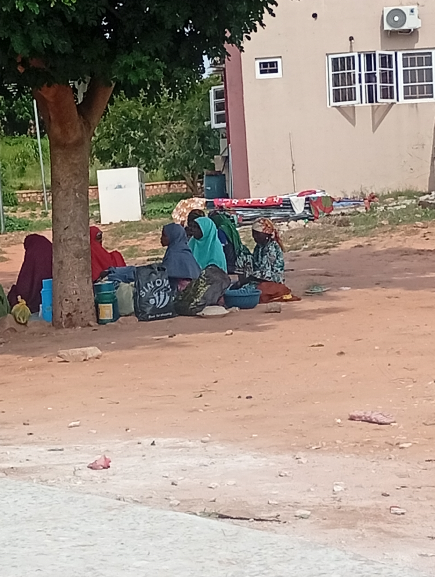 The image shows a group of five individuals, likely women and girls, sitting on the ground outdoors. They are gathered near the base of a large tree. The individuals are wearing traditional clothing, including hijabs and headscarves in various colors like maroon, red, blue, green, and patterned fabric. Some are seated directly on the sandy ground, while others have items like bags and possibly blankets around them.
One individual in the foreground, on the right, is seated with her back mostly to the camera, wearing a patterned green and black garment and an orange-brown headscarf. In front of her is a blue basin. Other visible objects include several bags, one black bag with white lettering that appears to say "SINO", and a green cylindrical container. A yellow bag is visible to the left, near the base of the tree.
The environment appears to be an outdoor area with a sandy or dirt ground. In the background, there is a light-colored building with windows and an air conditioning unit. There are also some scattered plants and a stone wall or structure. The overall scene suggests a casual gathering or resting spot, possibly in a public area or near a residence. The specific location in Gombe, Nigeria, is not identifiable from the visual cues alone; it could be a street, a community area, or near a commercial or residential building.