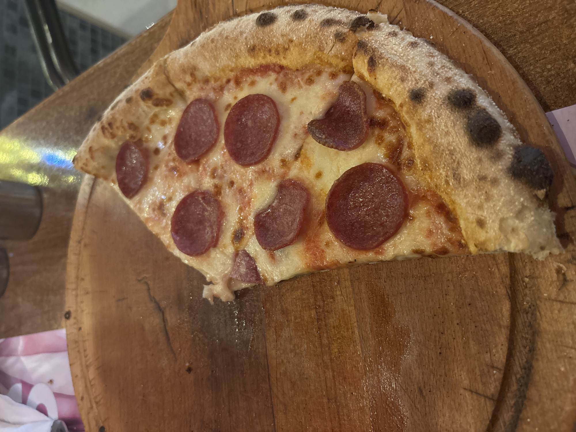A close-up, high-angle shot shows a slice of pepperoni pizza on a wooden board. The pizza has a golden-brown crust with some charred spots, melted white cheese, and several circular slices of pepperoni. The wooden board has a rough texture and some visible grain. In the background, out of focus, are hints of a pink and white paper napkin and a blurred yellow and blue light source. The scene appears to be indoors, possibly in a restaurant, and suggests a casual dining experience. Without further context or visual cues, it is impossible to determine the specific location within the city of Amman, Jordan.